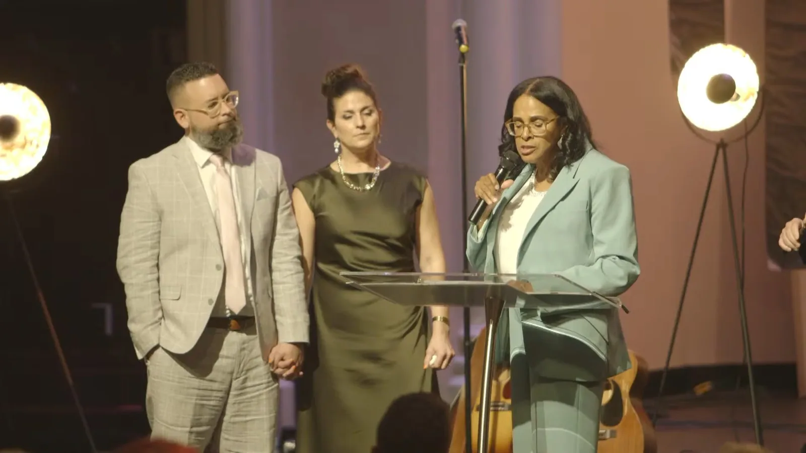 Two couples standing at the clear podium on stage, one woman speaking into a microphone