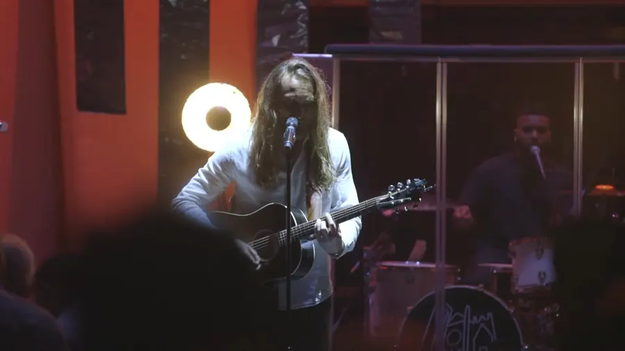 A worship leader with an acoustic guitar under warm stage light, shot over the heads of the crowd