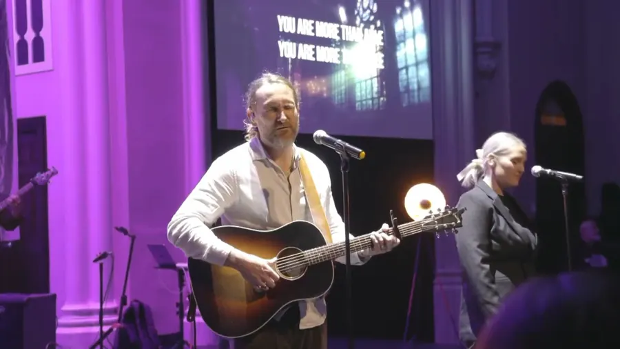 A worship leader with a guitar under purple light, with lyrics on the screen behind him