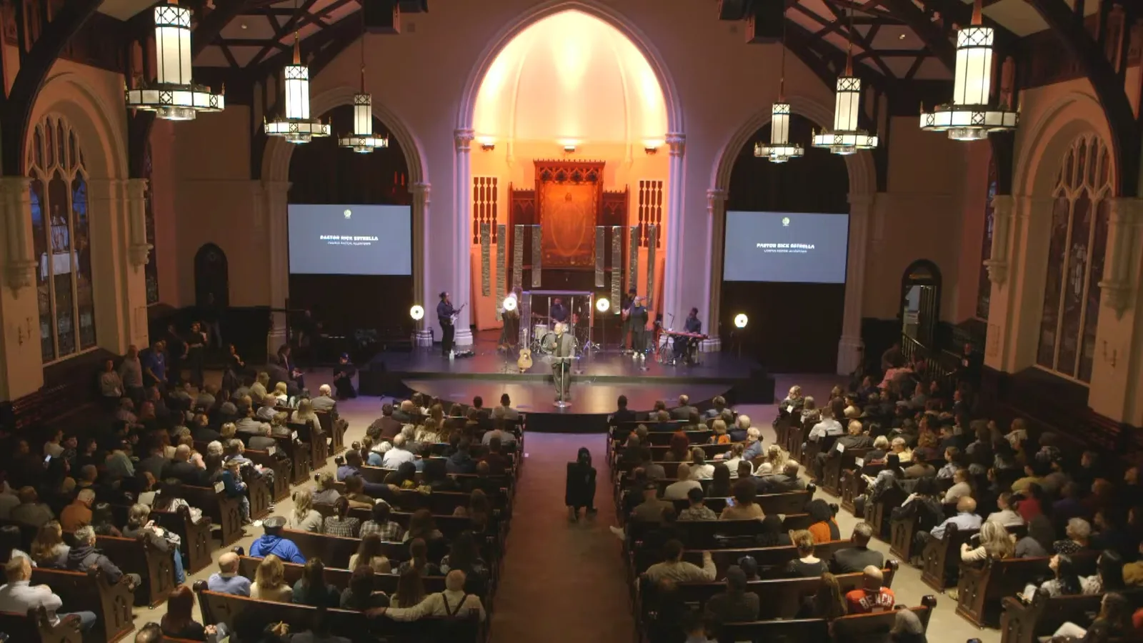 A packed sanctuary seen from high at the back, with pews full, two screens on either side of a lit arch and the band on stage