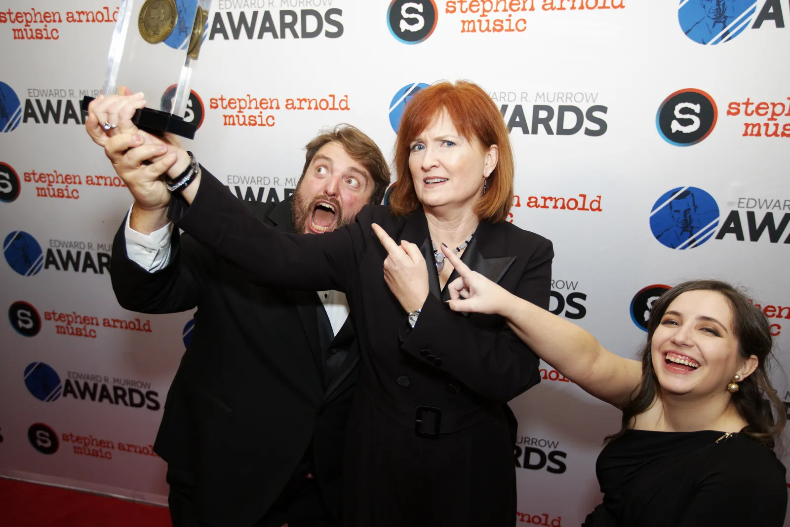 A man holding his award high over his head while a colleague points at it and another woman laughs beside them