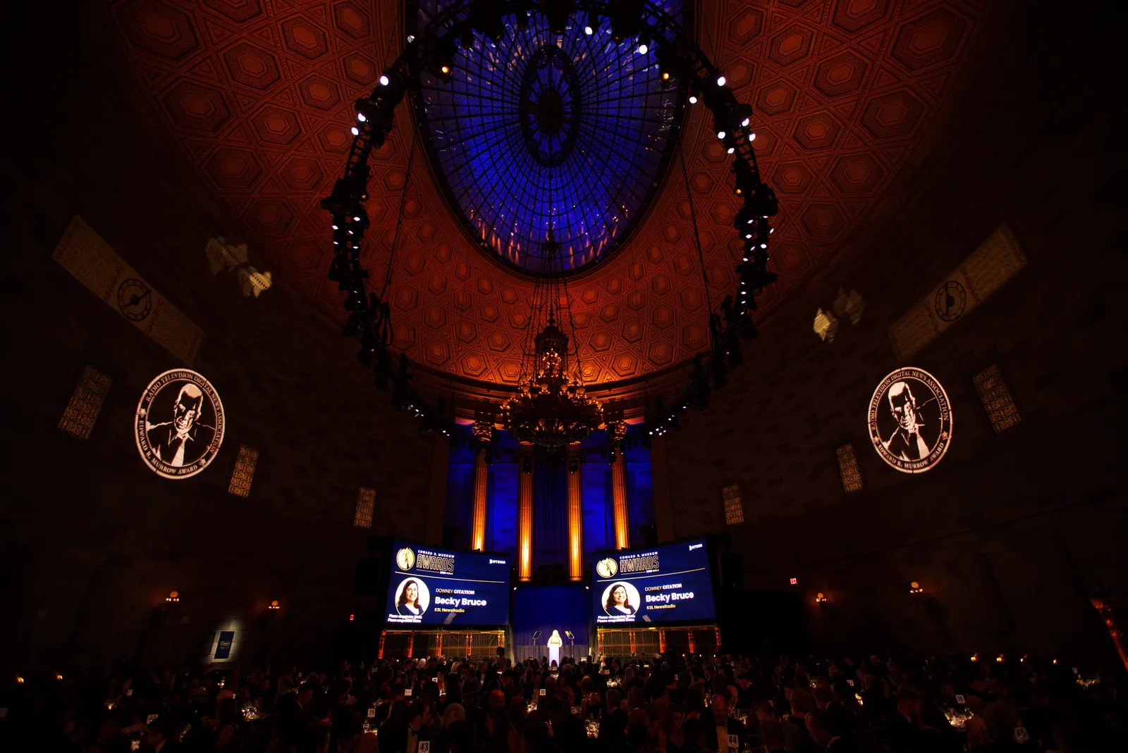 The Gotham Hall ballroom from the back of the room, with the lit dome ceiling, Murrow medallions on the walls and the stage screens in the distance