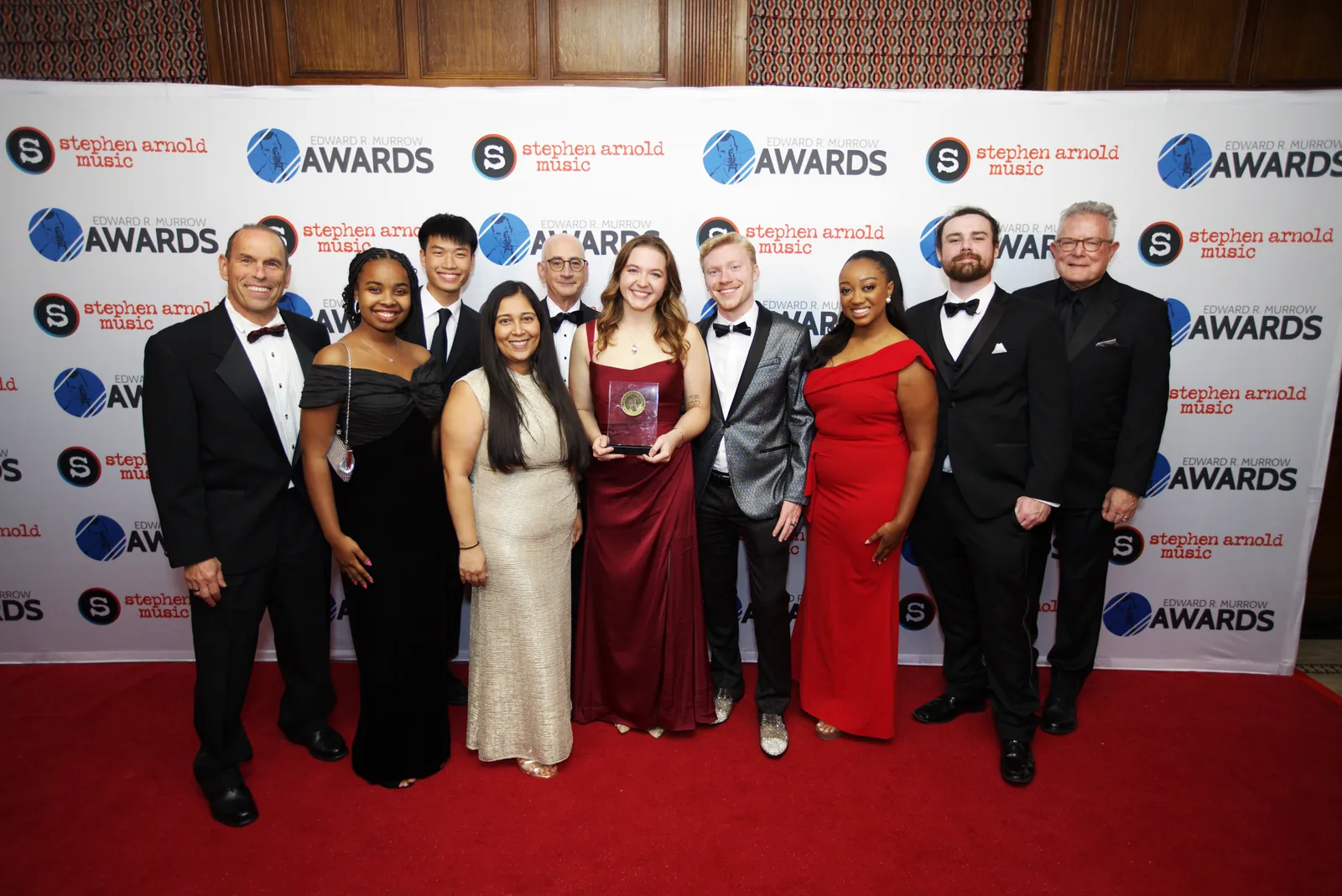 A group of about twelve journalists in formal wear lined up on the red carpet, one holding a Murrow Award