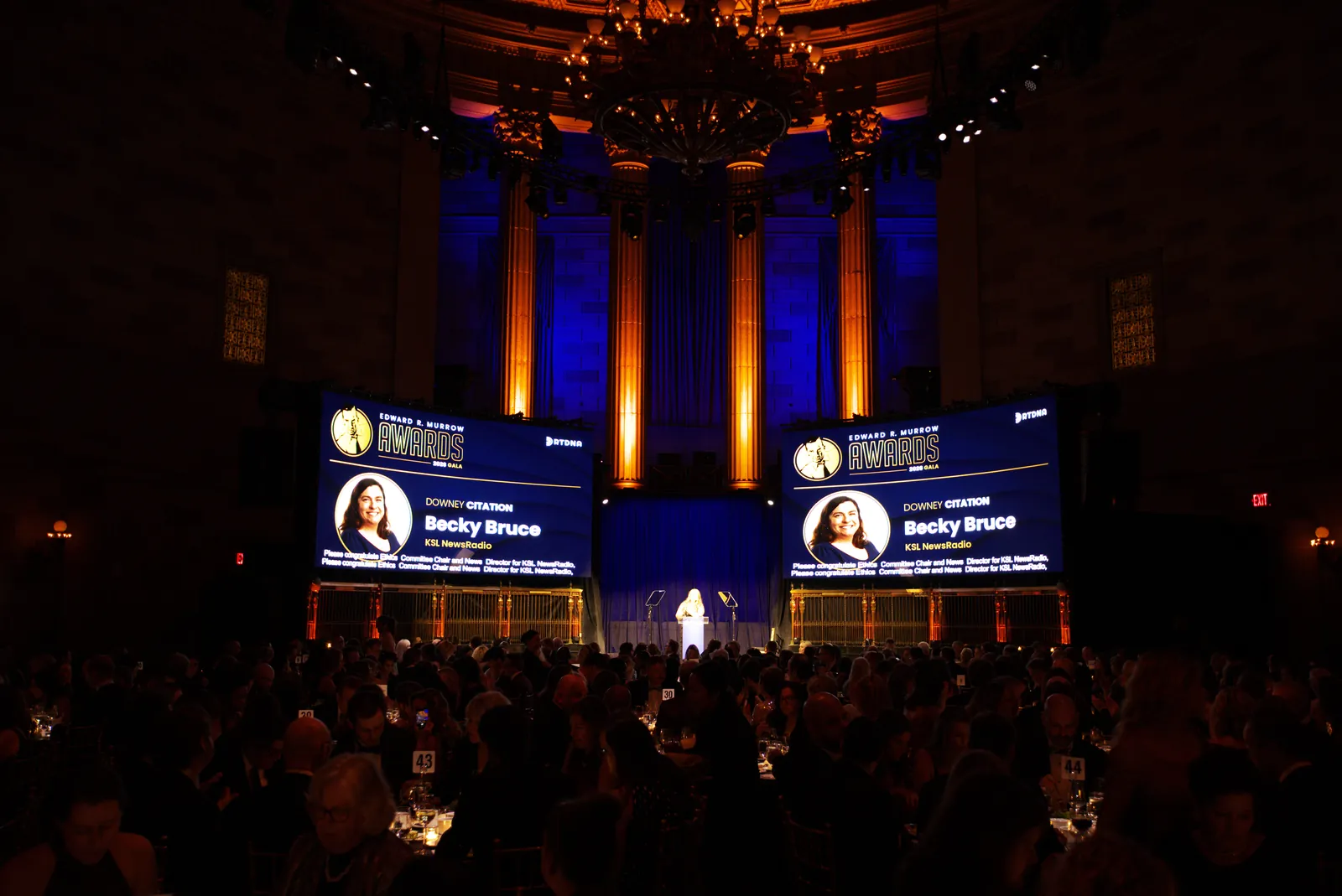 The main stage at Gotham Hall with two large screens showing an award recipient and blue light on the columns behind