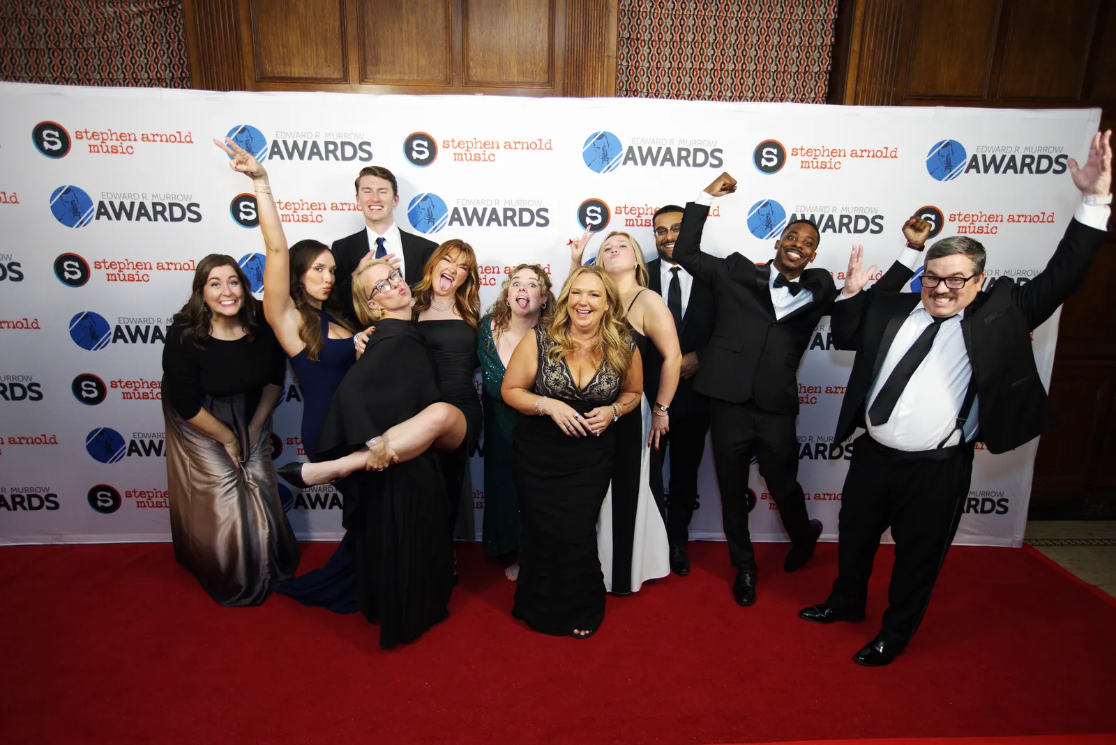 A large group in formal wear on the red carpet, several with arms in the air cheering