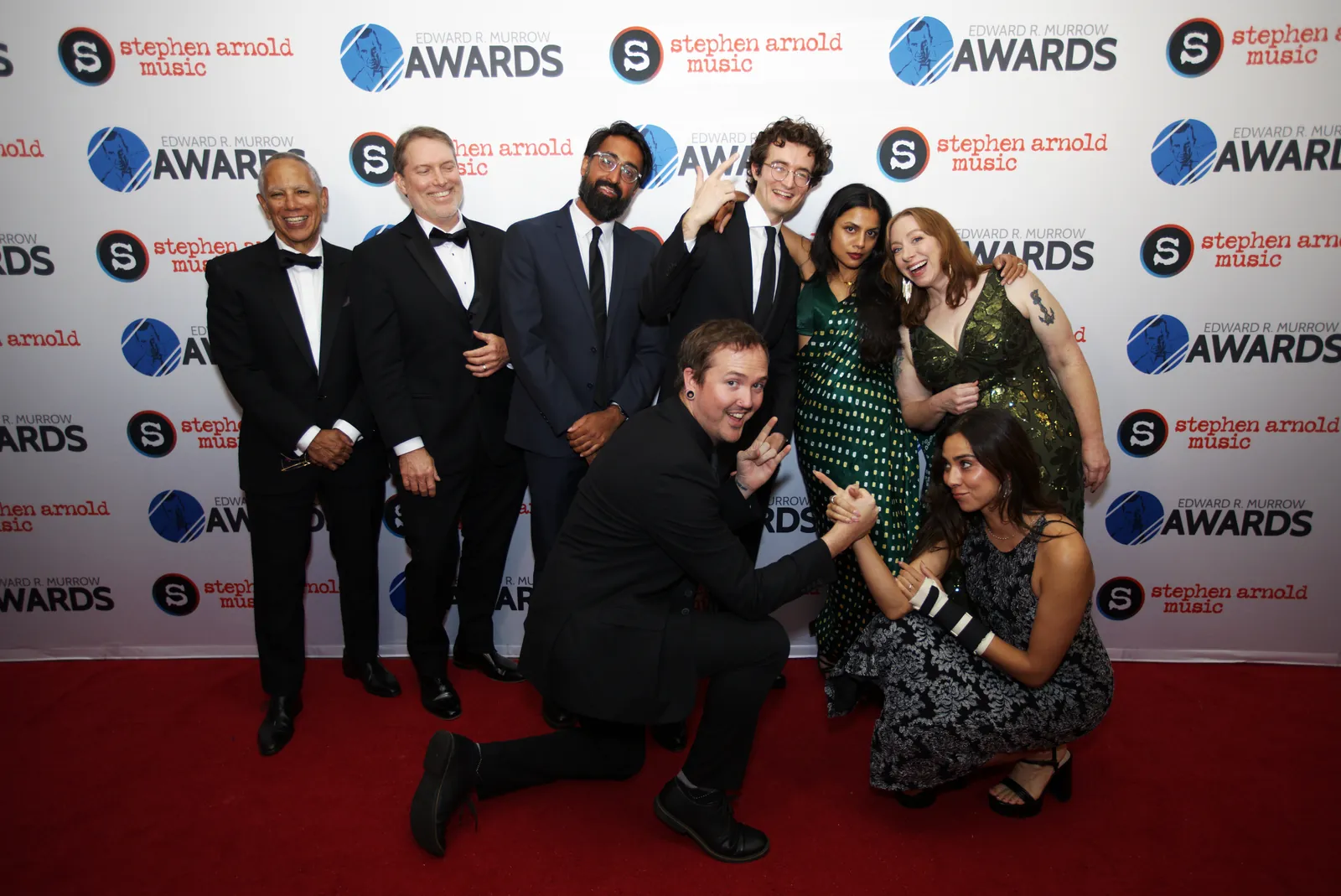 A newsroom team posing on the red carpet with one man kneeling in front and a woman crouched beside him