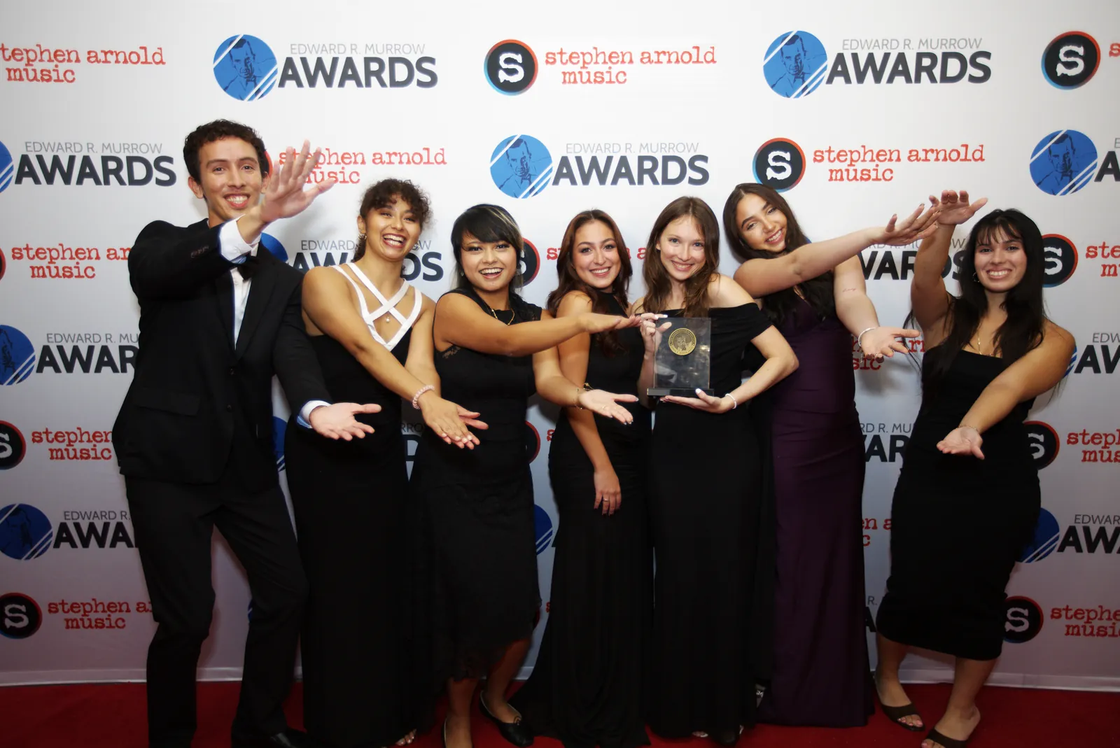 A team of eight in black formal wear on the red carpet, all pointing at the crystal award one of them is holding