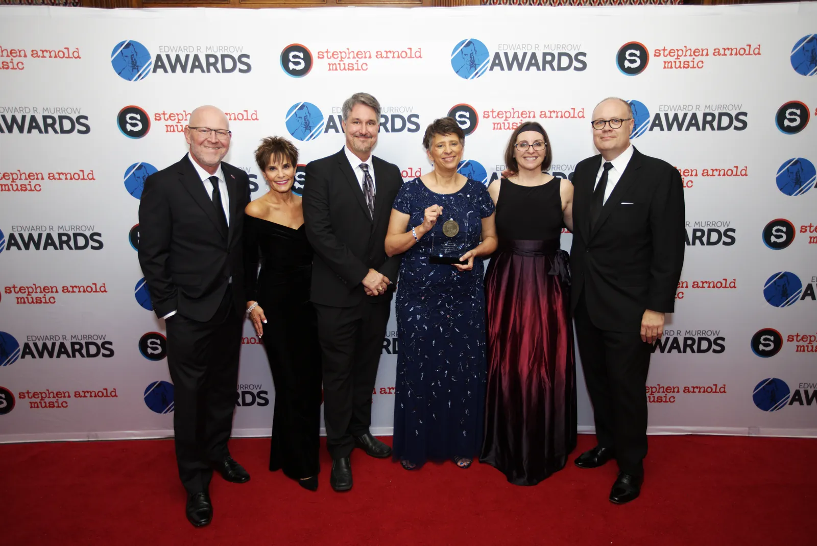 Six people in formal wear standing on the red carpet, one woman in a sequined blue gown holding an award