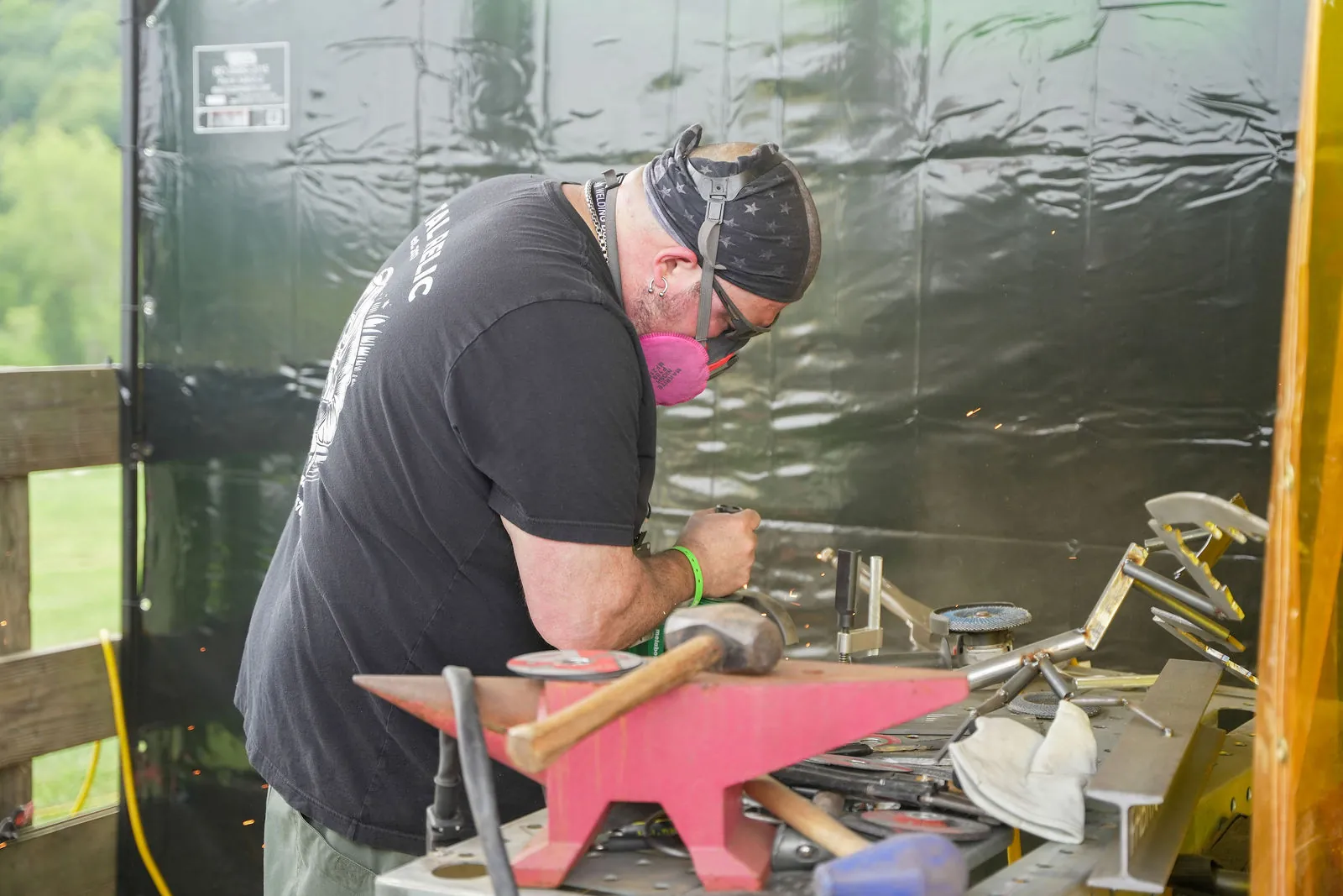 A man in a bandana and respirator shaping steel at a pink anvil in a work booth