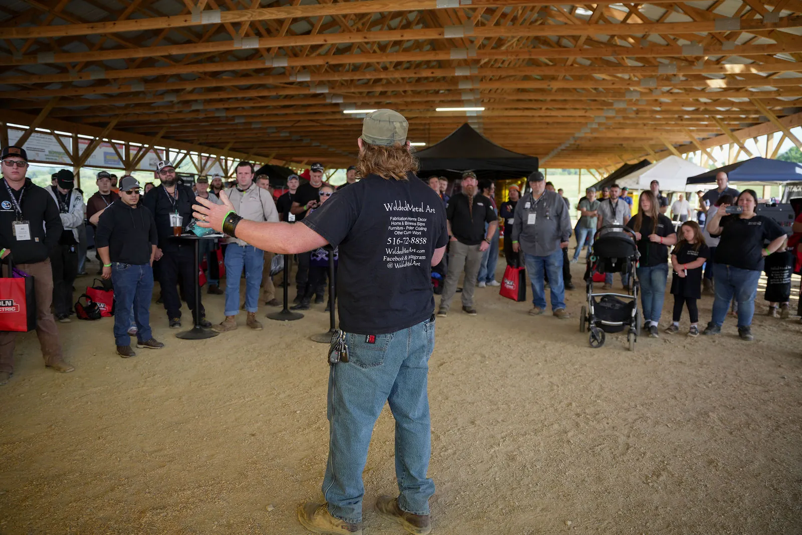A man in a Welded Metal Art shirt with his arms out, talking to a crowd gathered under the pavilion
