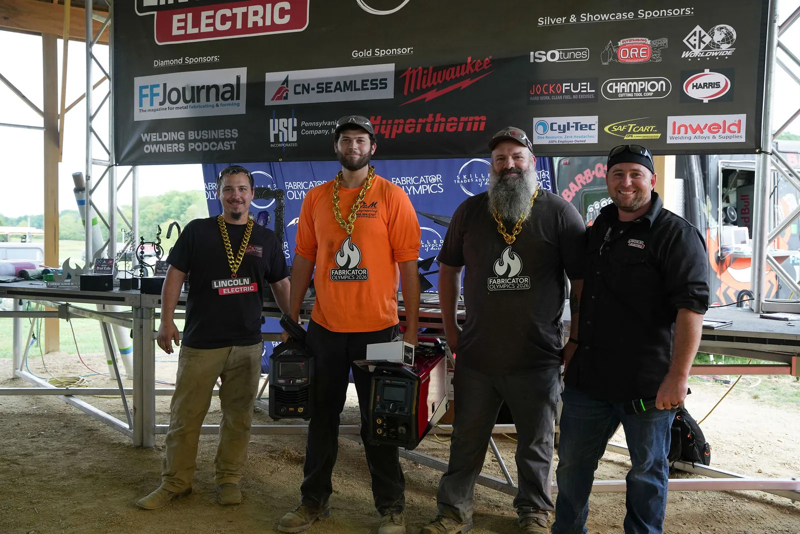 Four men on the main stage, three of them wearing gold chains, standing behind Lincoln Electric welders with the sponsor banner overhead