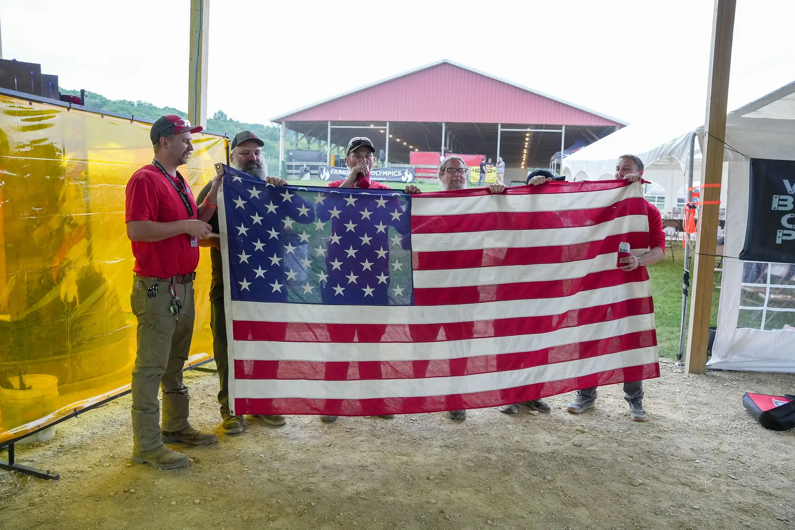 A group of people holding a large American flag between yellow welding curtains, with the red-roofed grandstand of the Kempton Fairgrounds behind them