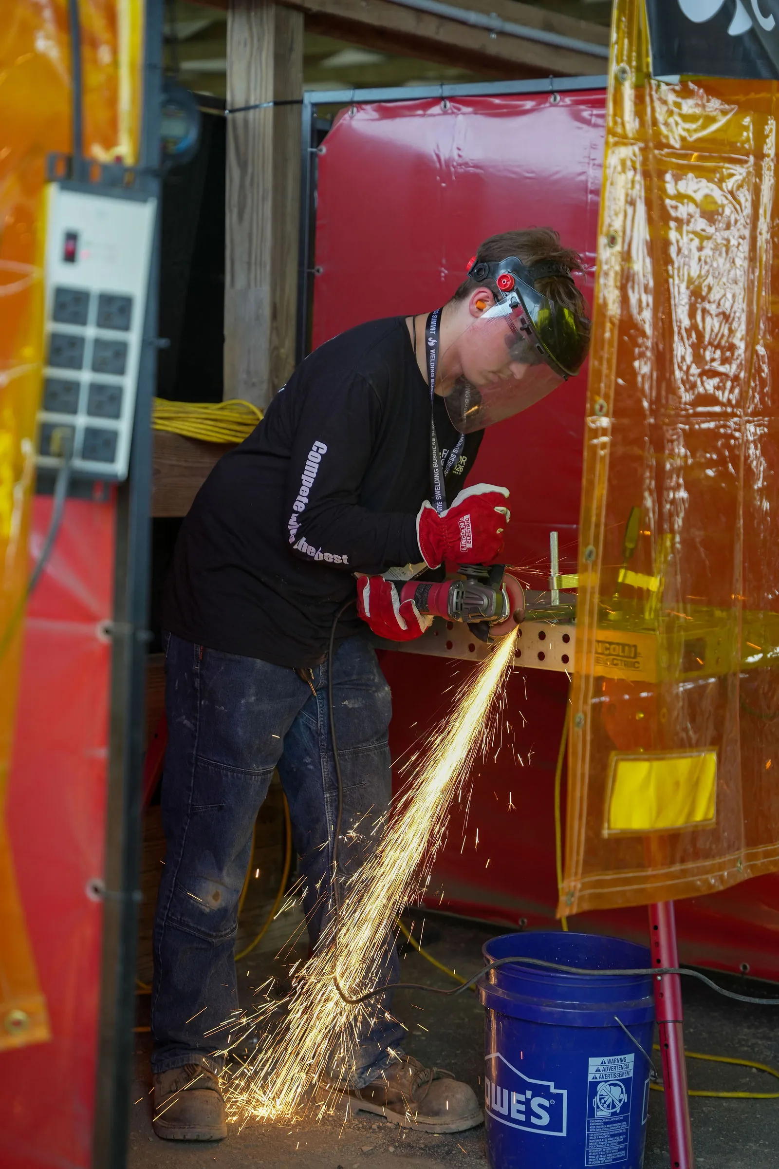 A man grinding steel inside a booth with a stream of sparks falling toward a bucket on the ground