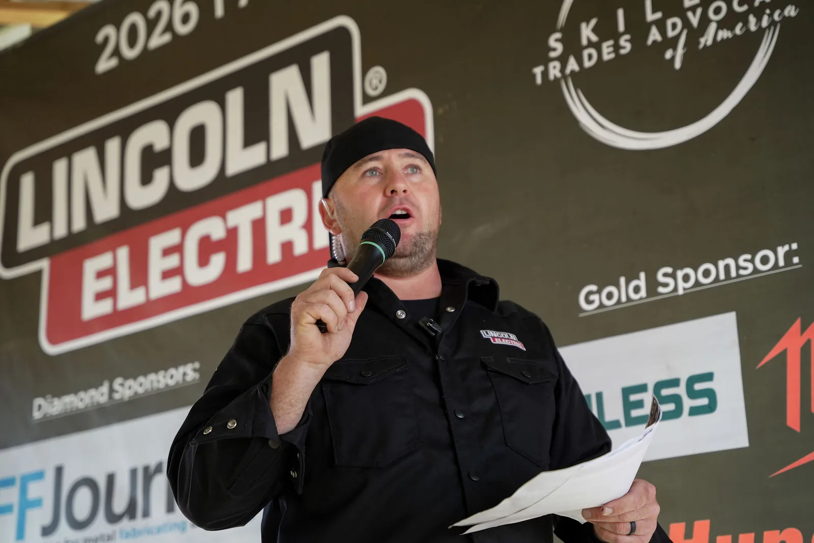 A man in a black cap and black work shirt speaking into a microphone in front of a Lincoln Electric sponsor banner
