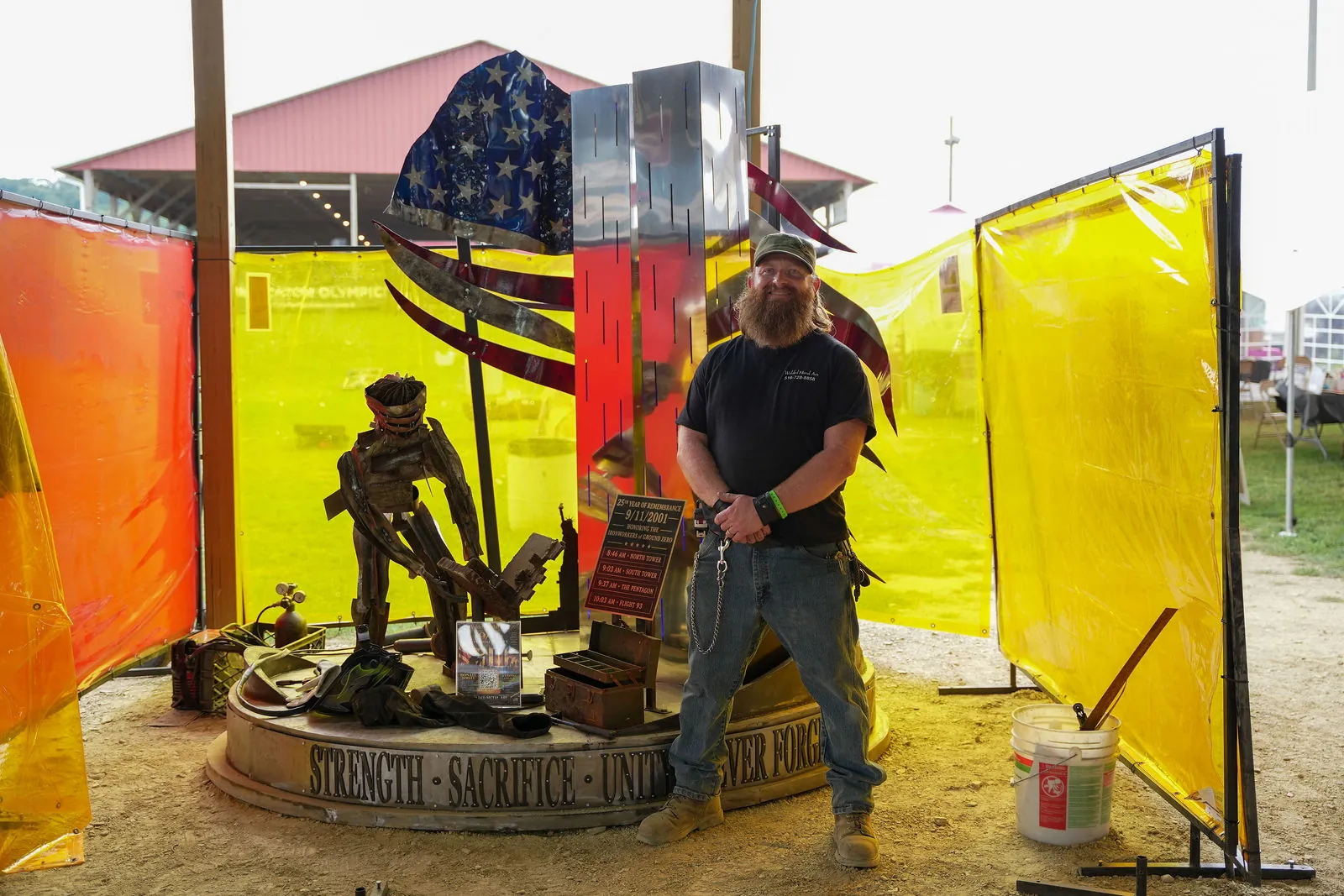 A large metal 9/11 memorial sculpture with a steel American flag and the words Strength, Sacrifice, Unity and Never Forget around the base, with a bearded man standing beside it between yellow welding curtains