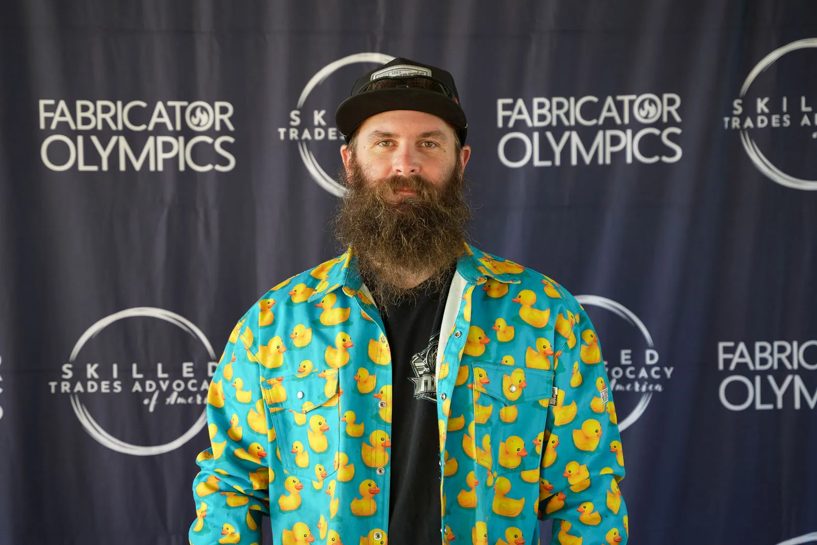 A bearded competitor in a black cap and a bright rubber duck print shirt in front of the Fabricator Olympics backdrop