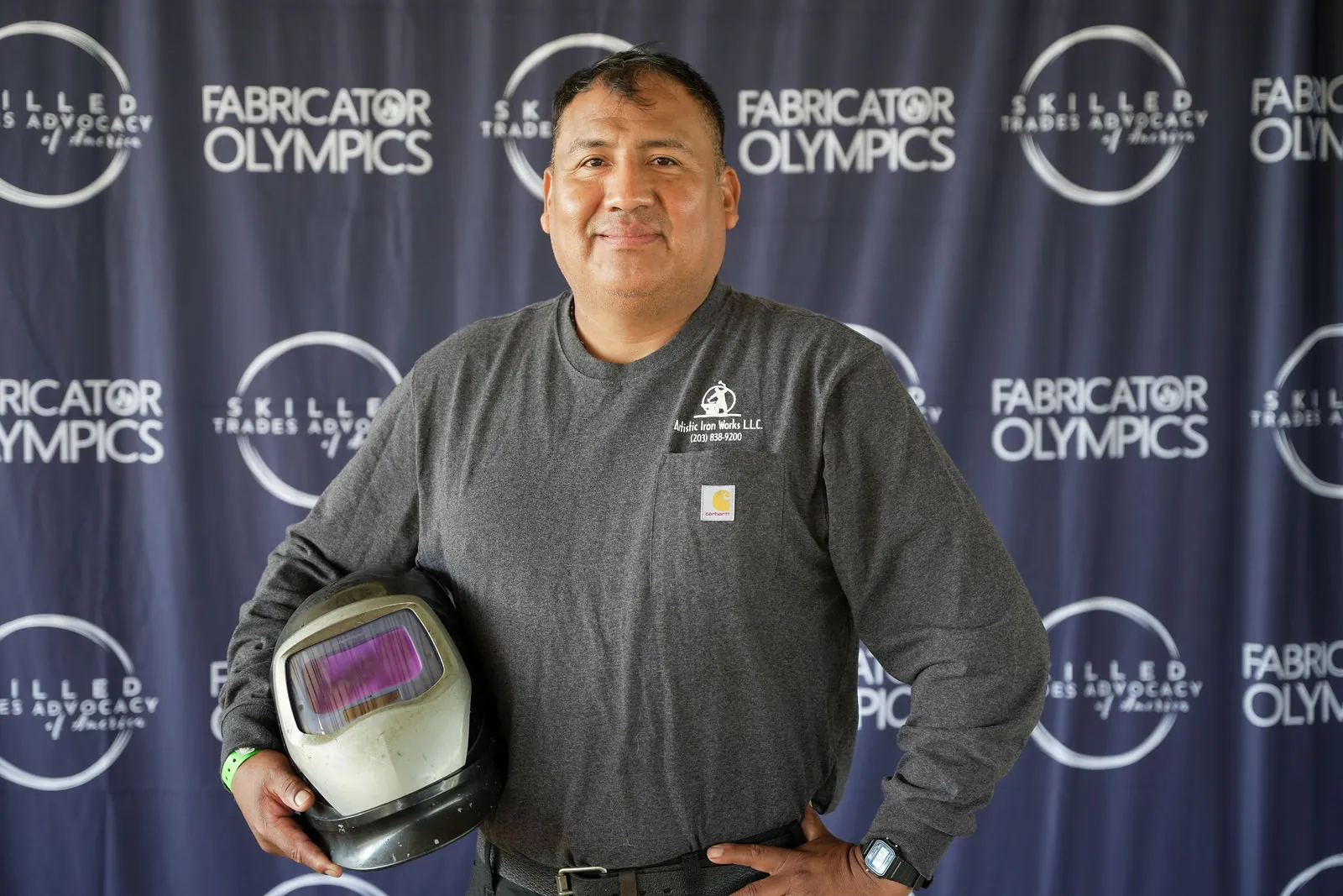 A competitor holding a welding helmet under his arm in front of the Fabricator Olympics backdrop