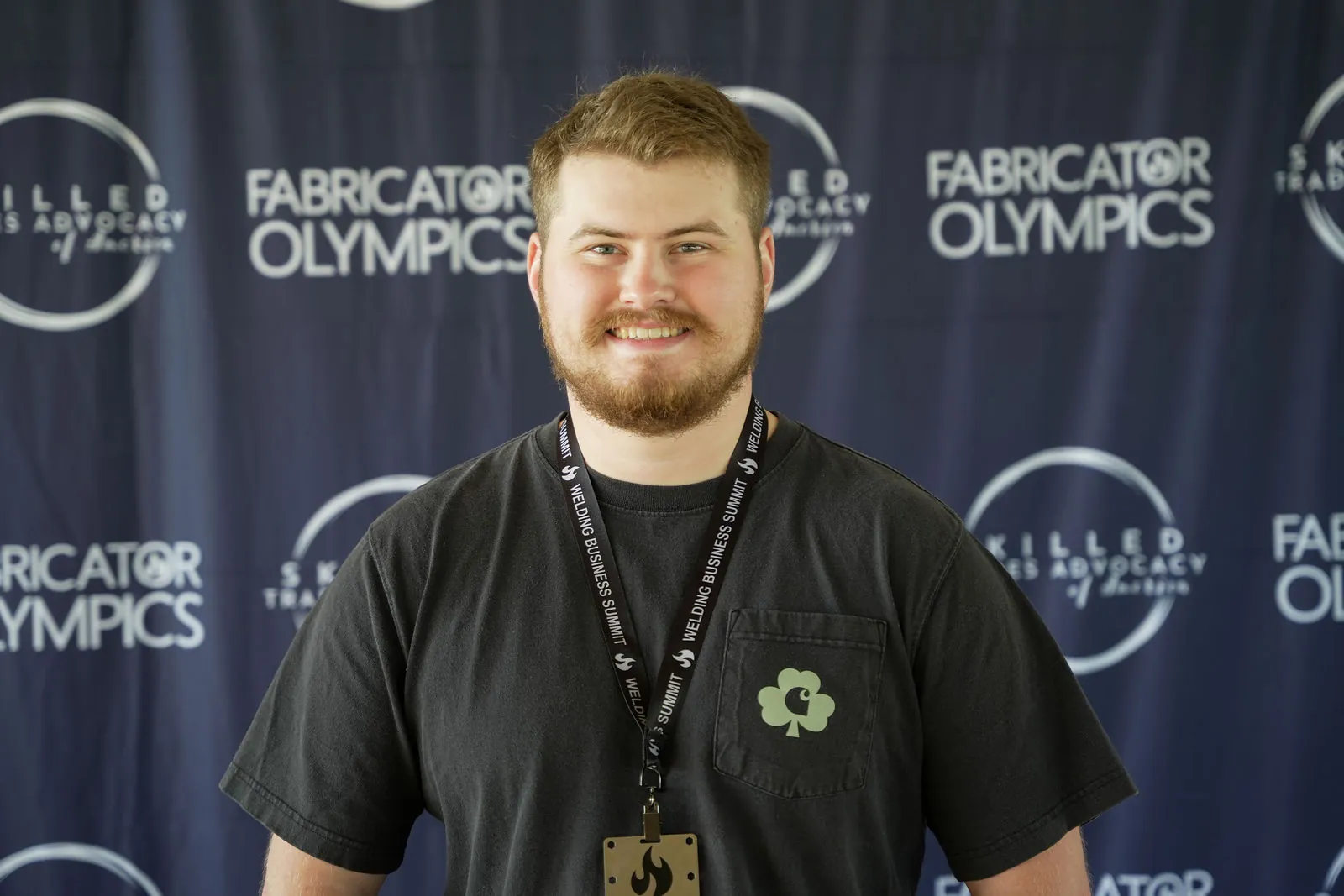 A young bearded competitor in a black shirt and lanyard in front of the Fabricator Olympics backdrop