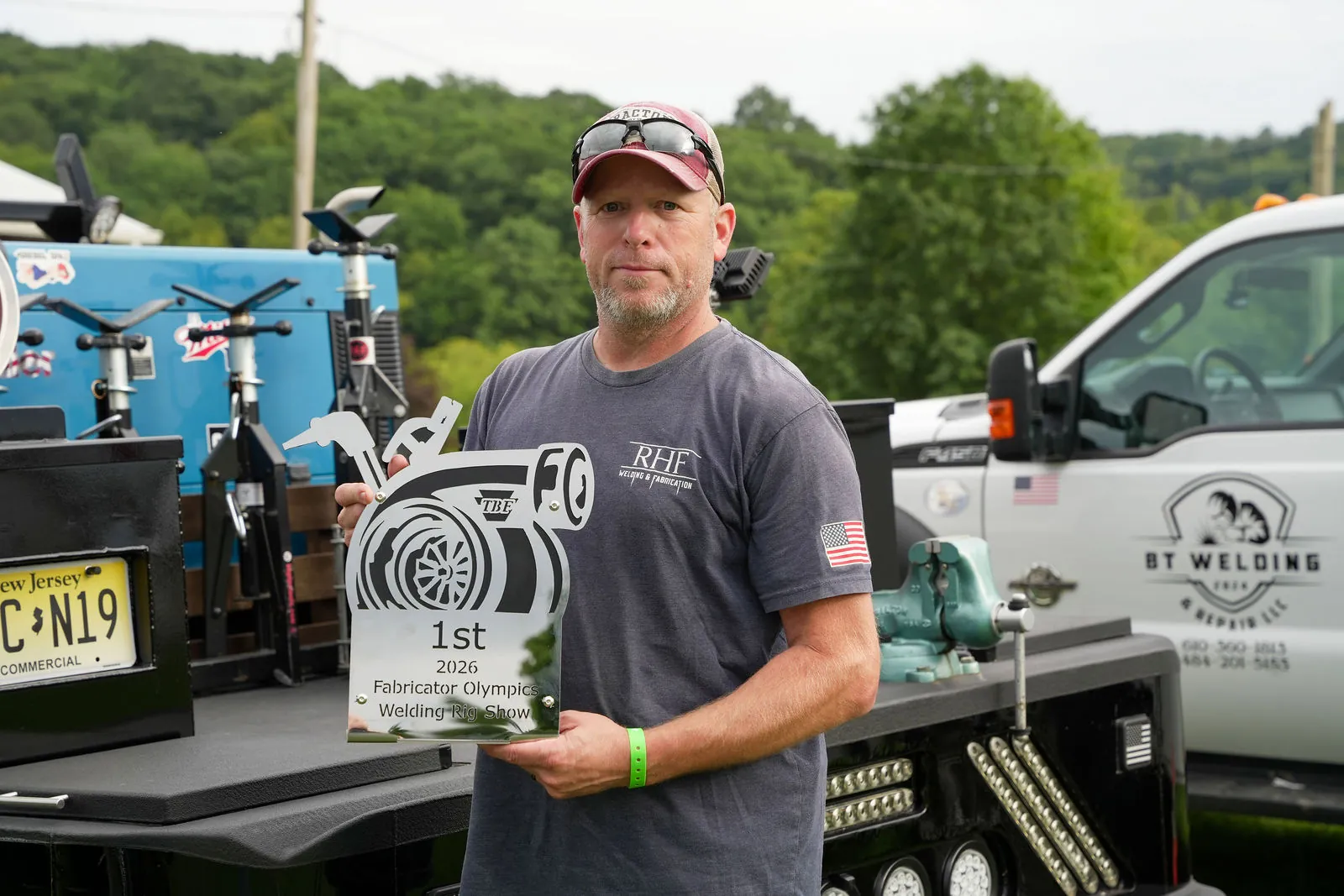 A man in a gray RHF Welding and Fabrication shirt holding a first place 2026 Fabricator Olympics rig show trophy in front of a welding rig