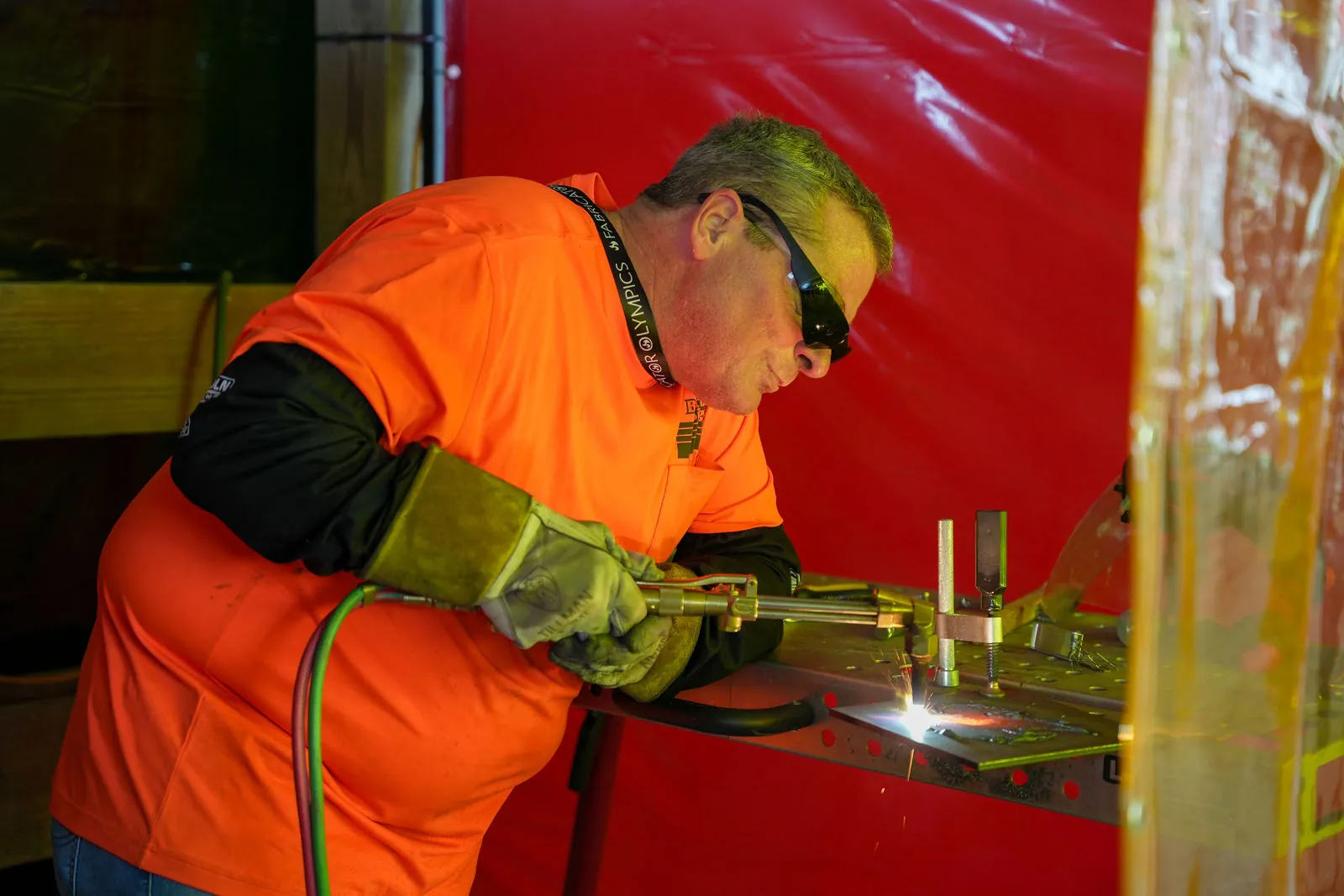 A man in an orange shirt, safety glasses and leather gloves cutting a piece of steel with a torch in front of a red curtain