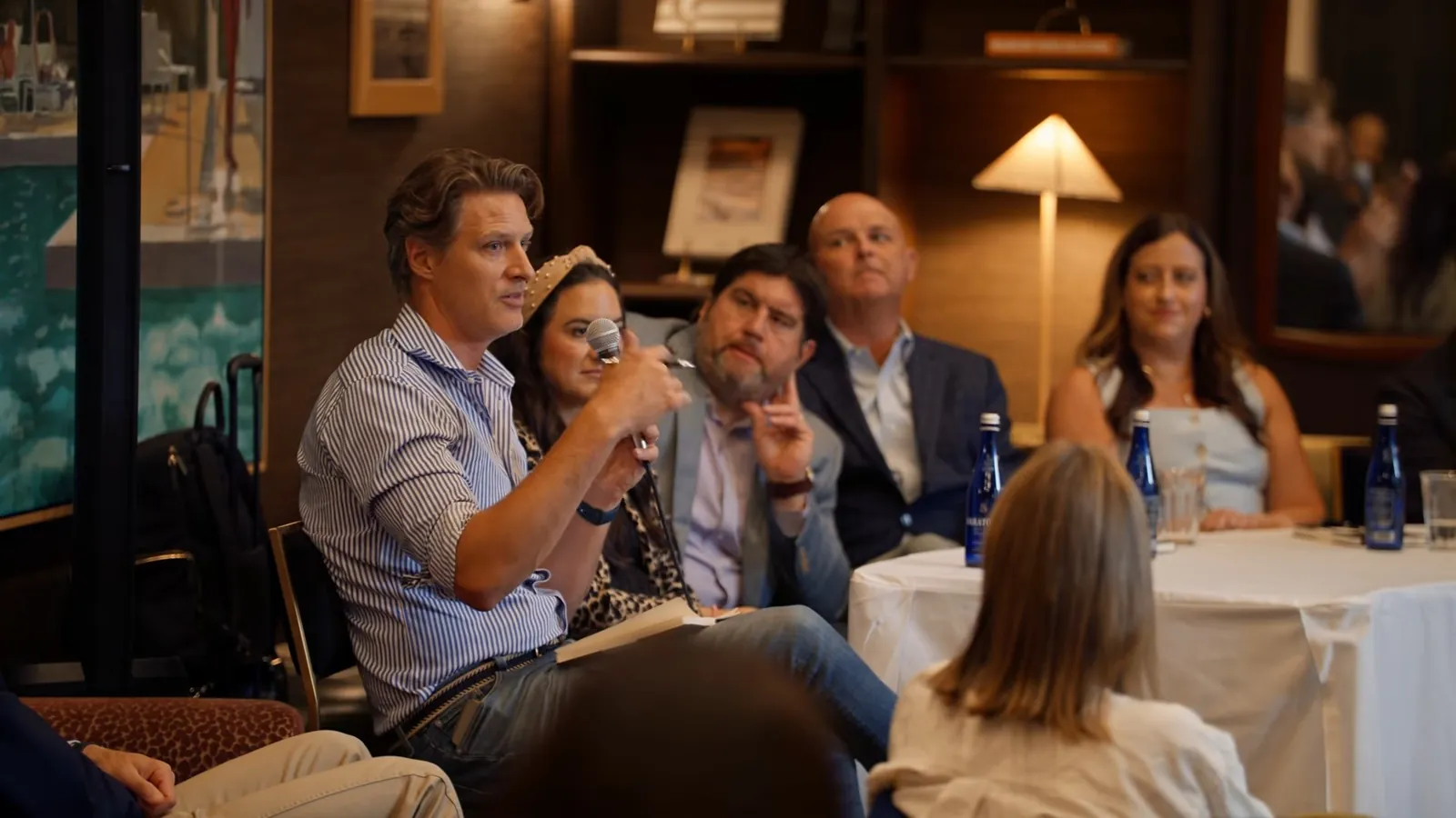 A panelist in a striped shirt speaking into a microphone, with other panelists listening beside him