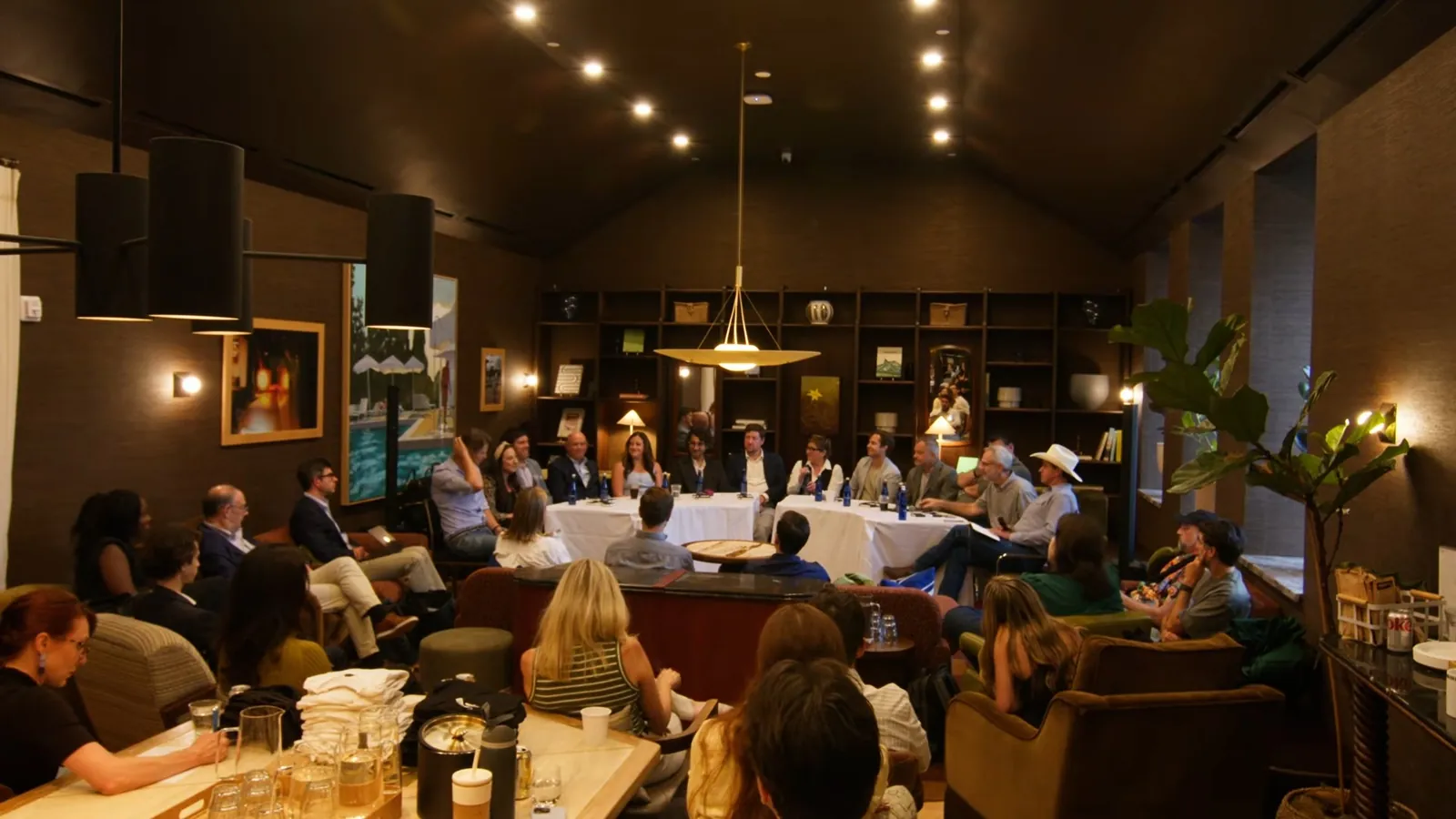 The full room from the back, with audience members in armchairs and couches and the panel at two tables at the far end