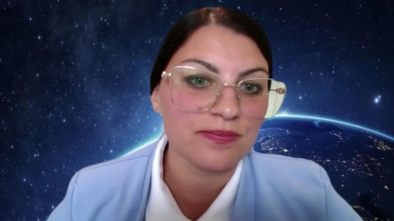 An interviewer in gold rimmed glasses and a light blue blazer on a video call with a space background