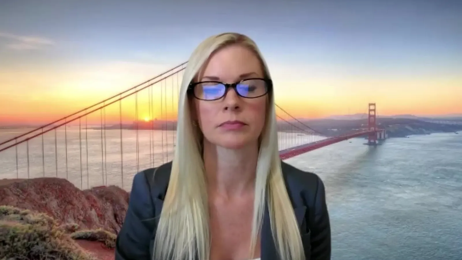 An interviewer with long blonde hair and glasses in a dark blazer on a video call with a bridge at sunset behind her