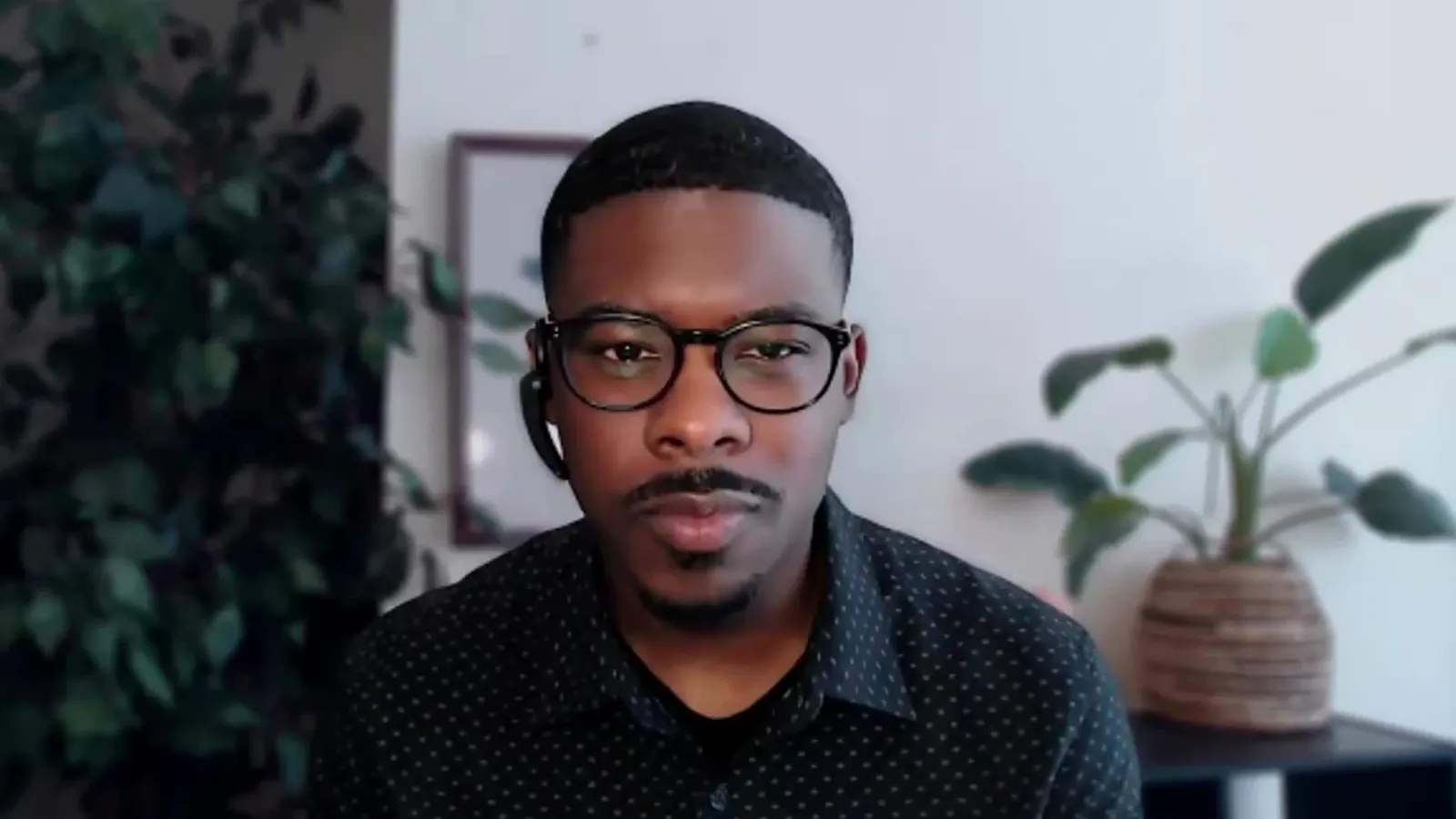 An interviewer in glasses and a dark patterned shirt on a video call, with plants and a framed print behind him