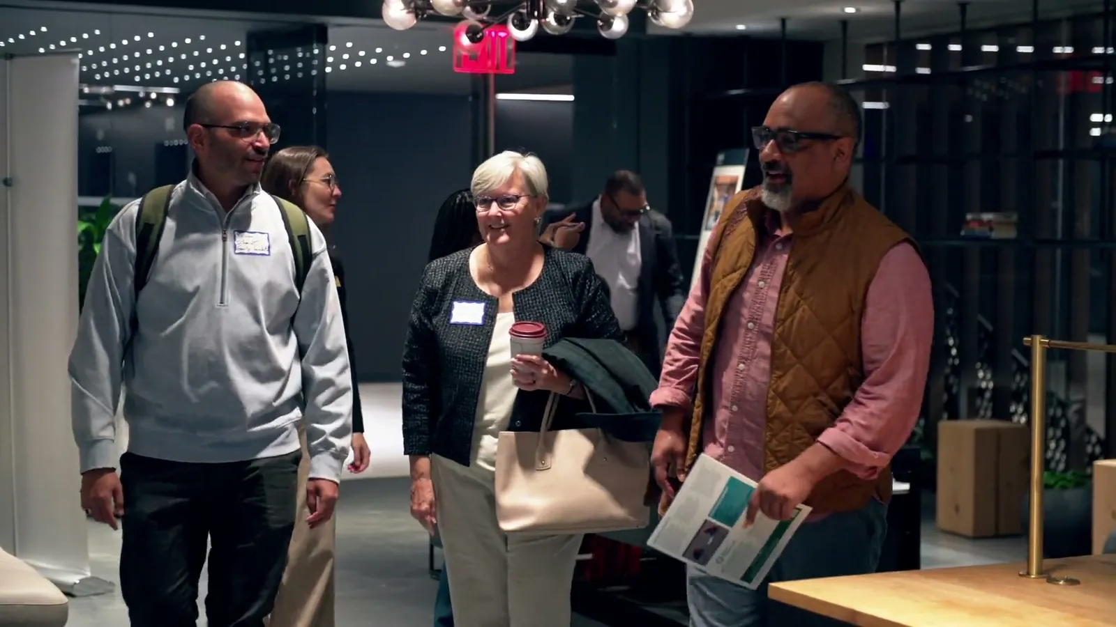 Three guests walking in with name tags, one holding a coffee and one carrying a program