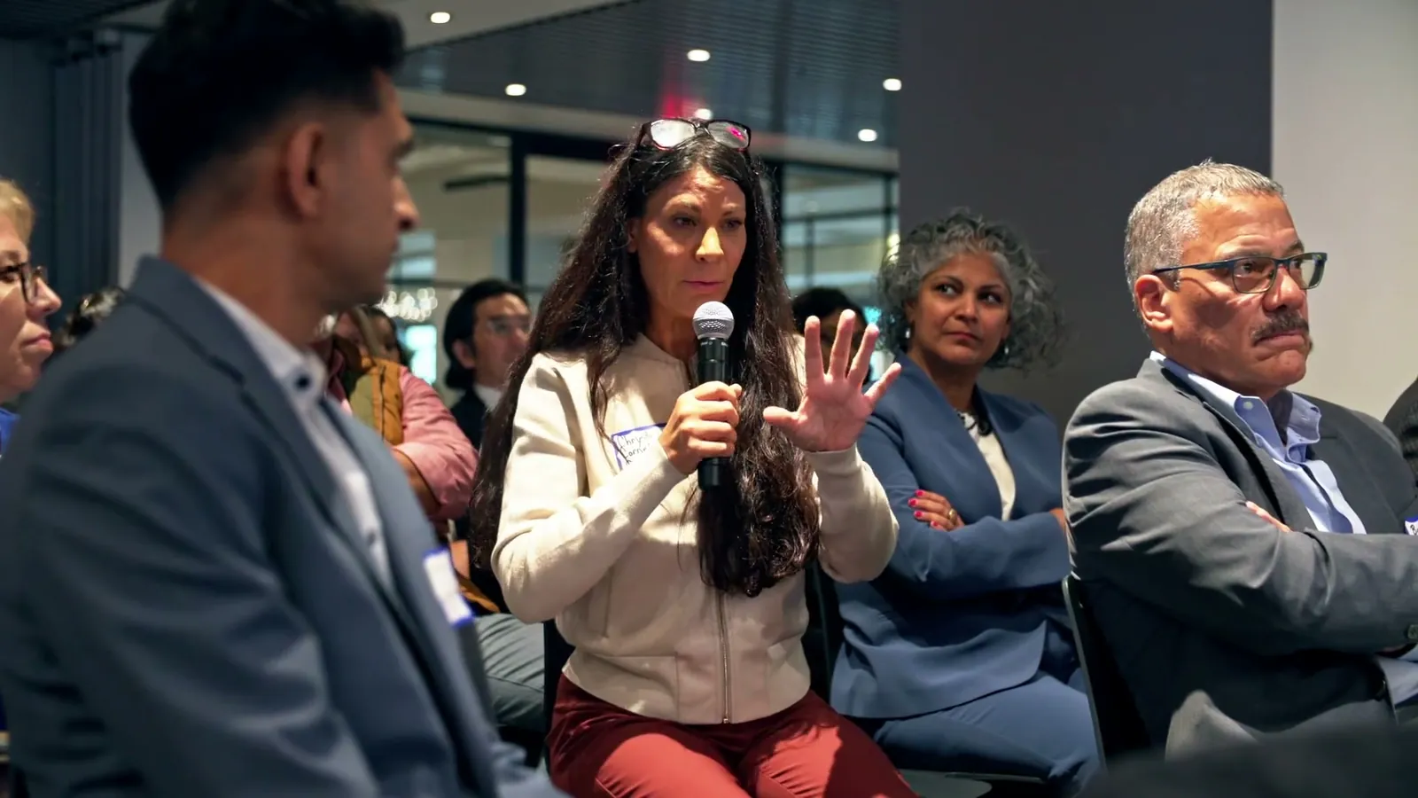 A woman in the audience holding a microphone and gesturing as she asks a question, with other guests seated around her
