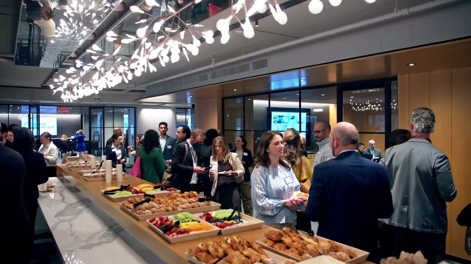 A long breakfast counter with pastries and fruit under hanging lights, with guests lined up along it