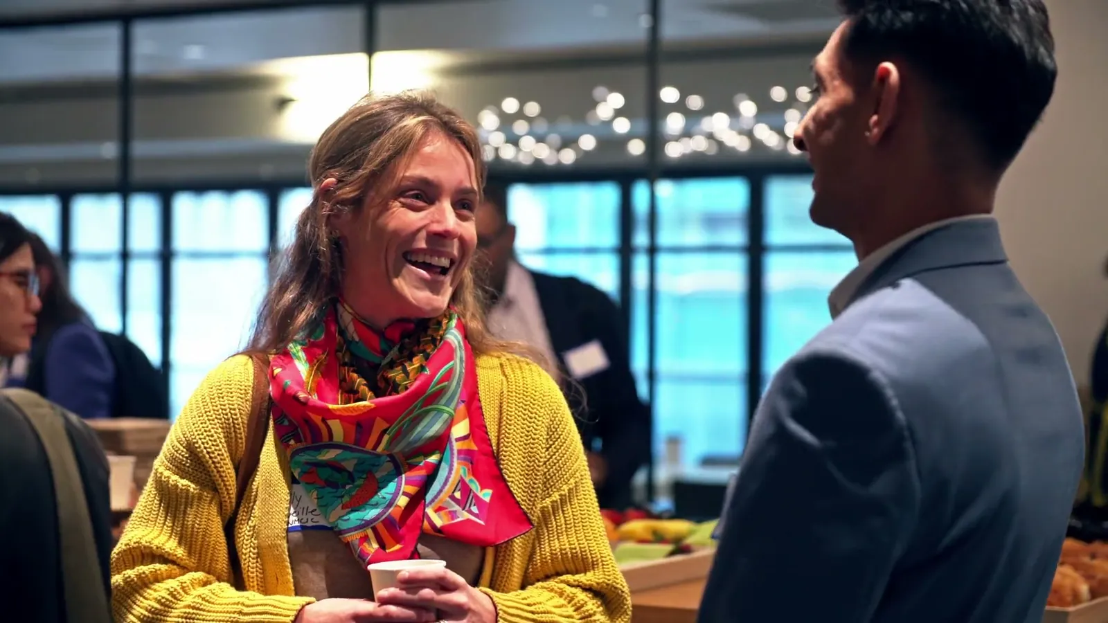 A woman in a yellow sweater and bright scarf laughing while she talks with a man in a suit