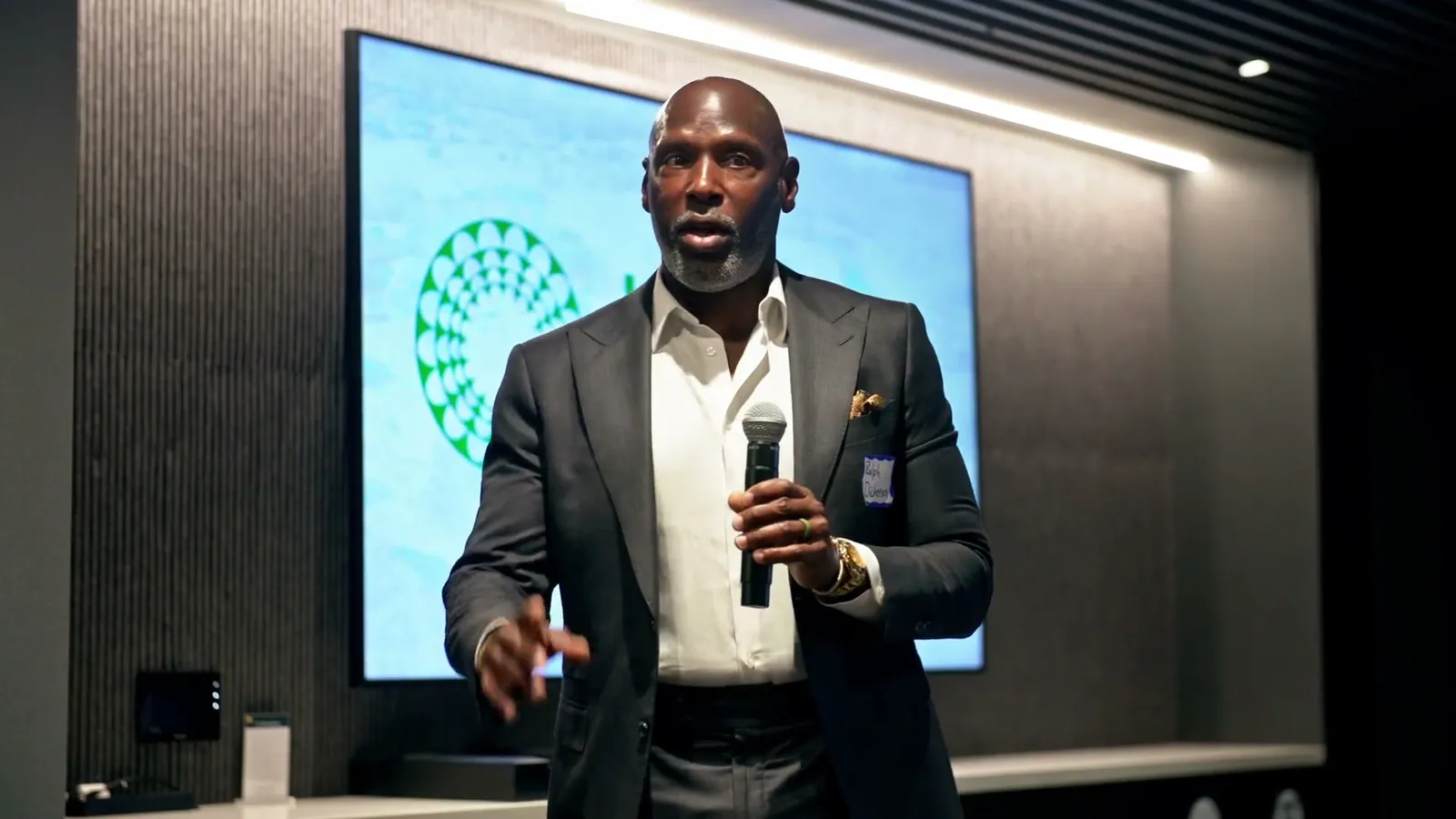 A speaker in a dark suit holding a microphone in front of a screen with the Justice Climate Fund logo