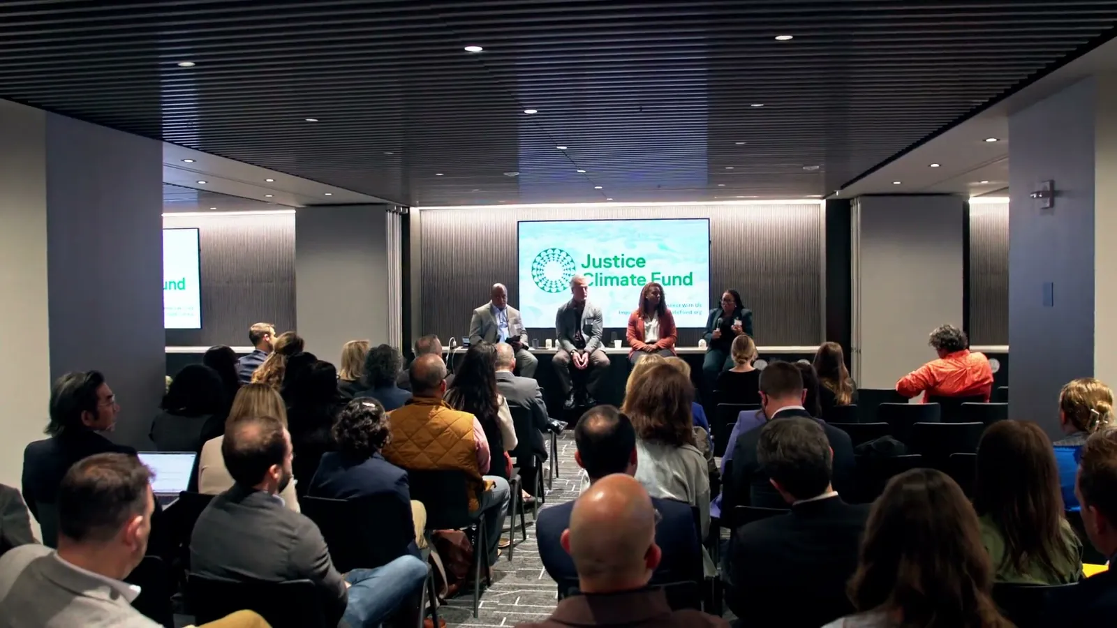 The panel seen from the back of the room over rows of seated guests, with the Justice Climate Fund logo on the screen