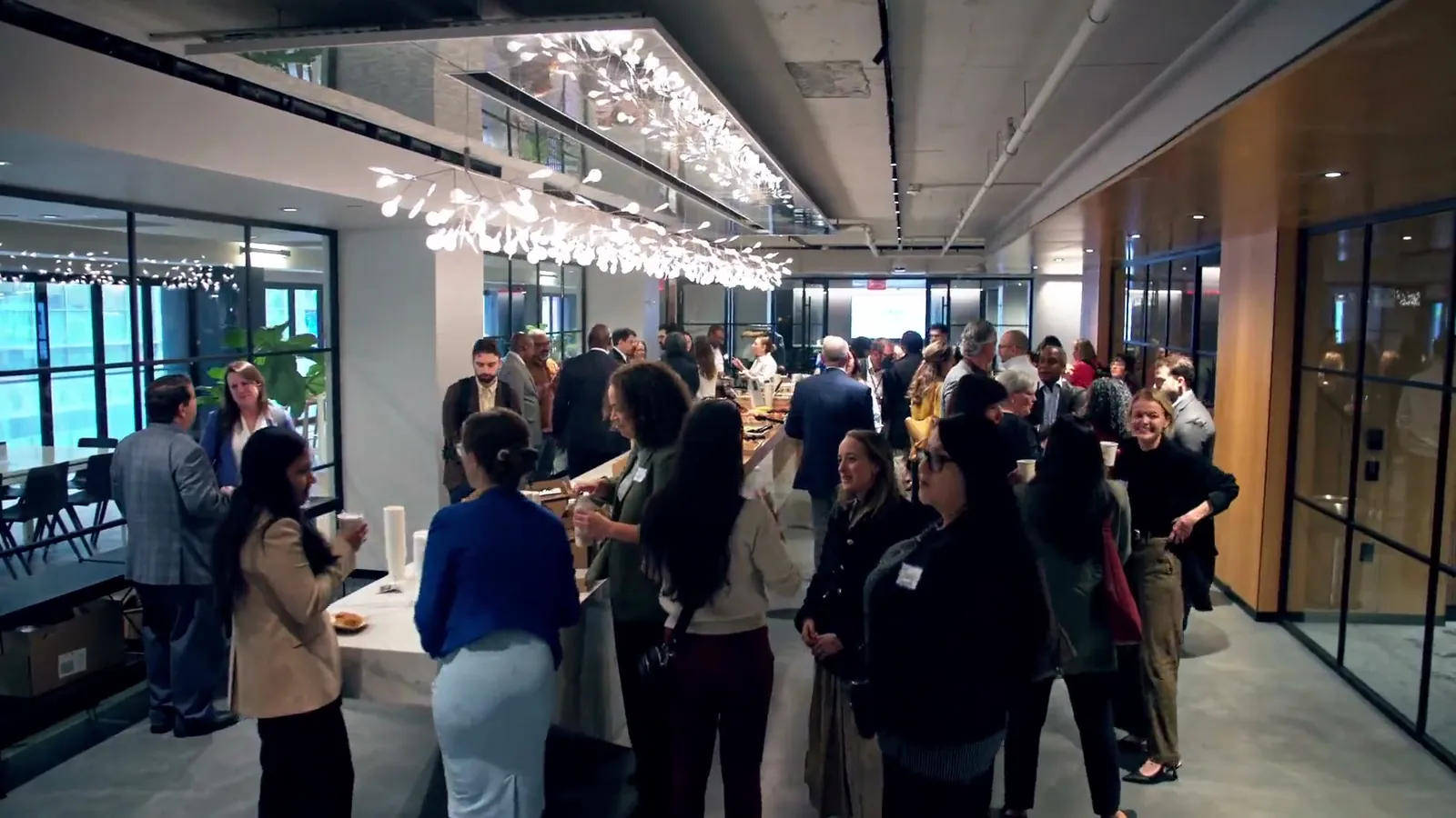 A crowded room of guests standing and talking, with tall windows along one side