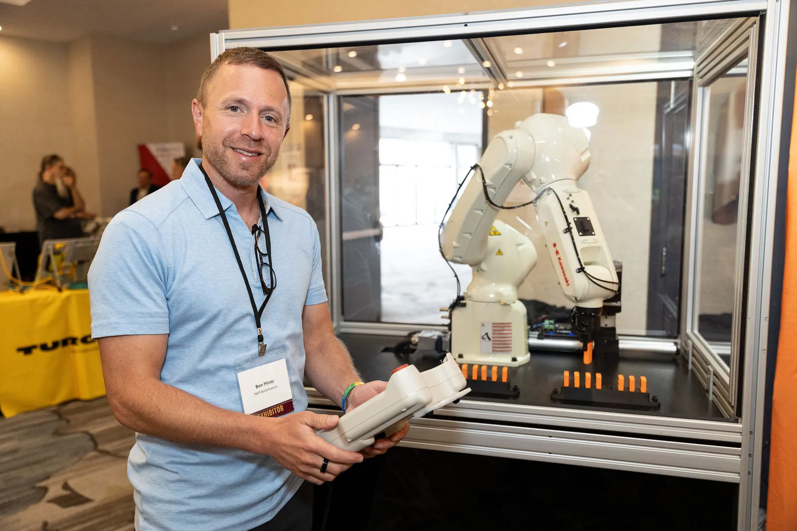 An exhibitor holding a robot teach pendant in front of a robot cell