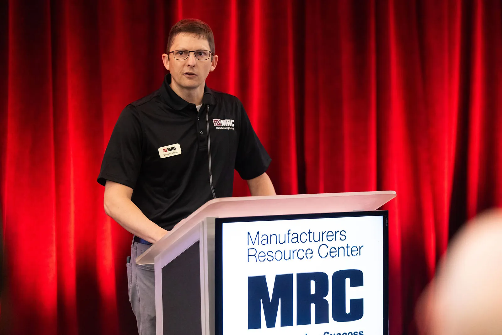 MRC's Shawn Furman speaking at the MRC podium in front of red curtains