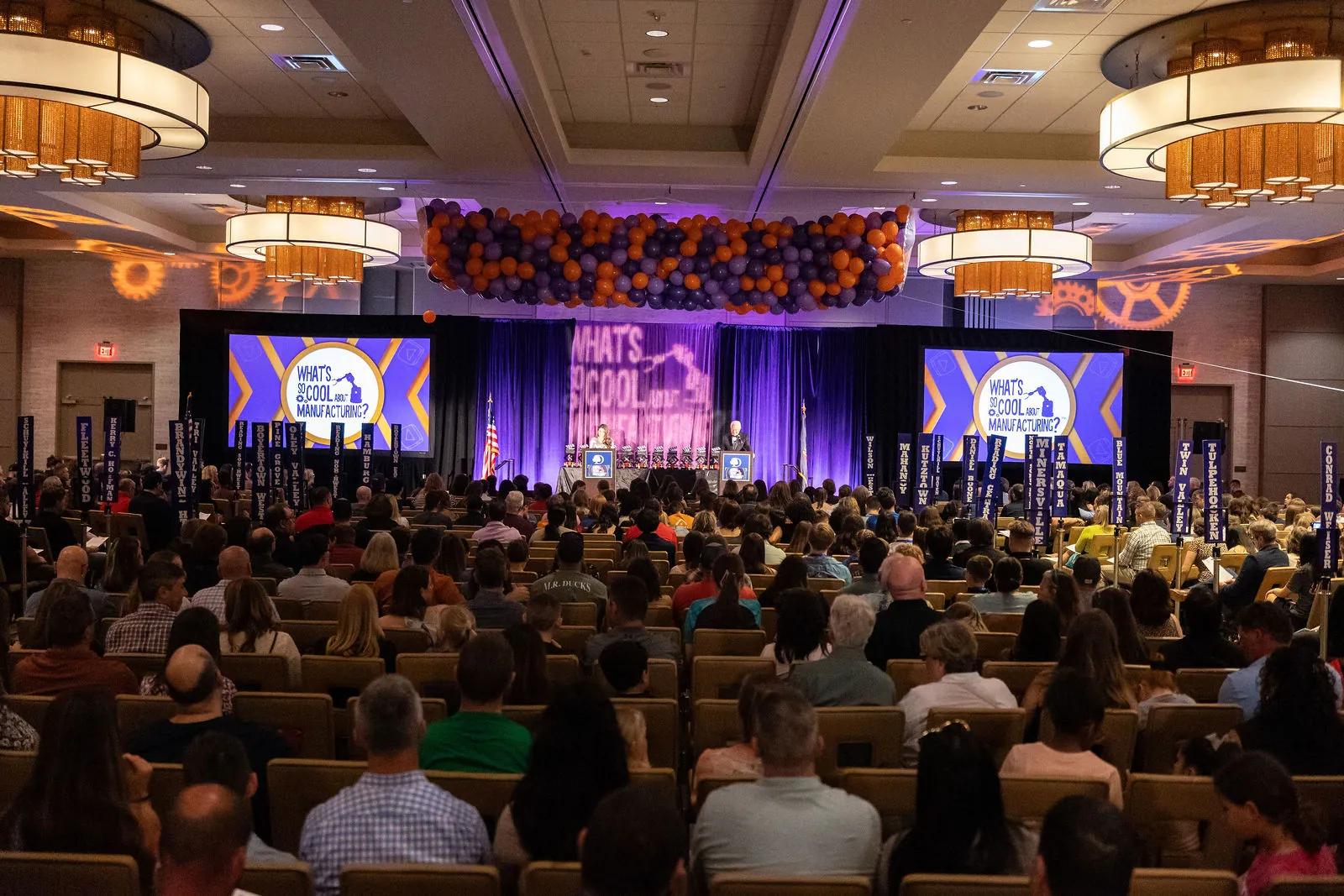 A packed hotel ballroom seen from the back of the room, with the contest logo on screens on both sides of the stage and purple and orange balloons hanging from the ceiling