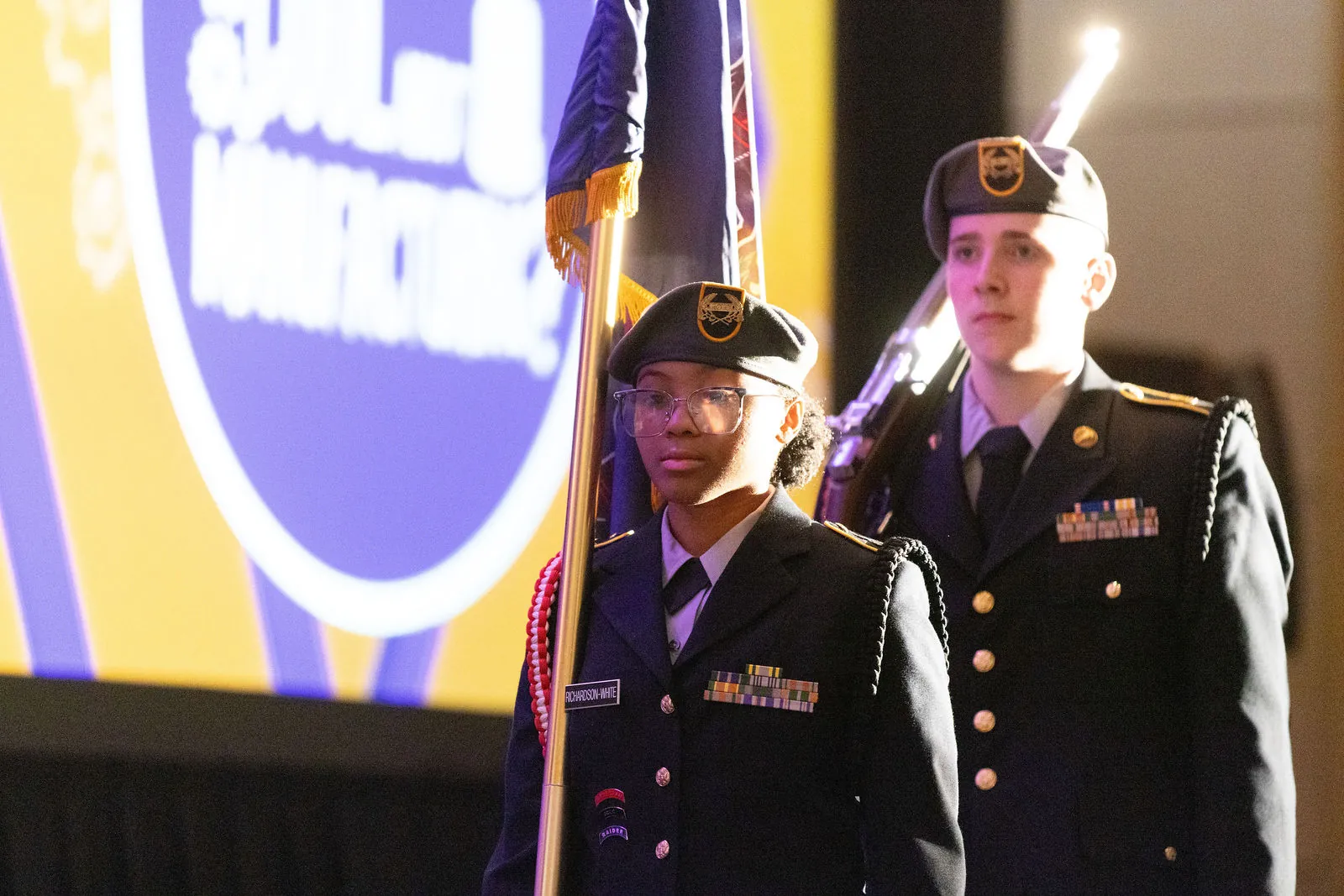Two students in dress uniforms carrying flags in front of the contest logo on a large screen