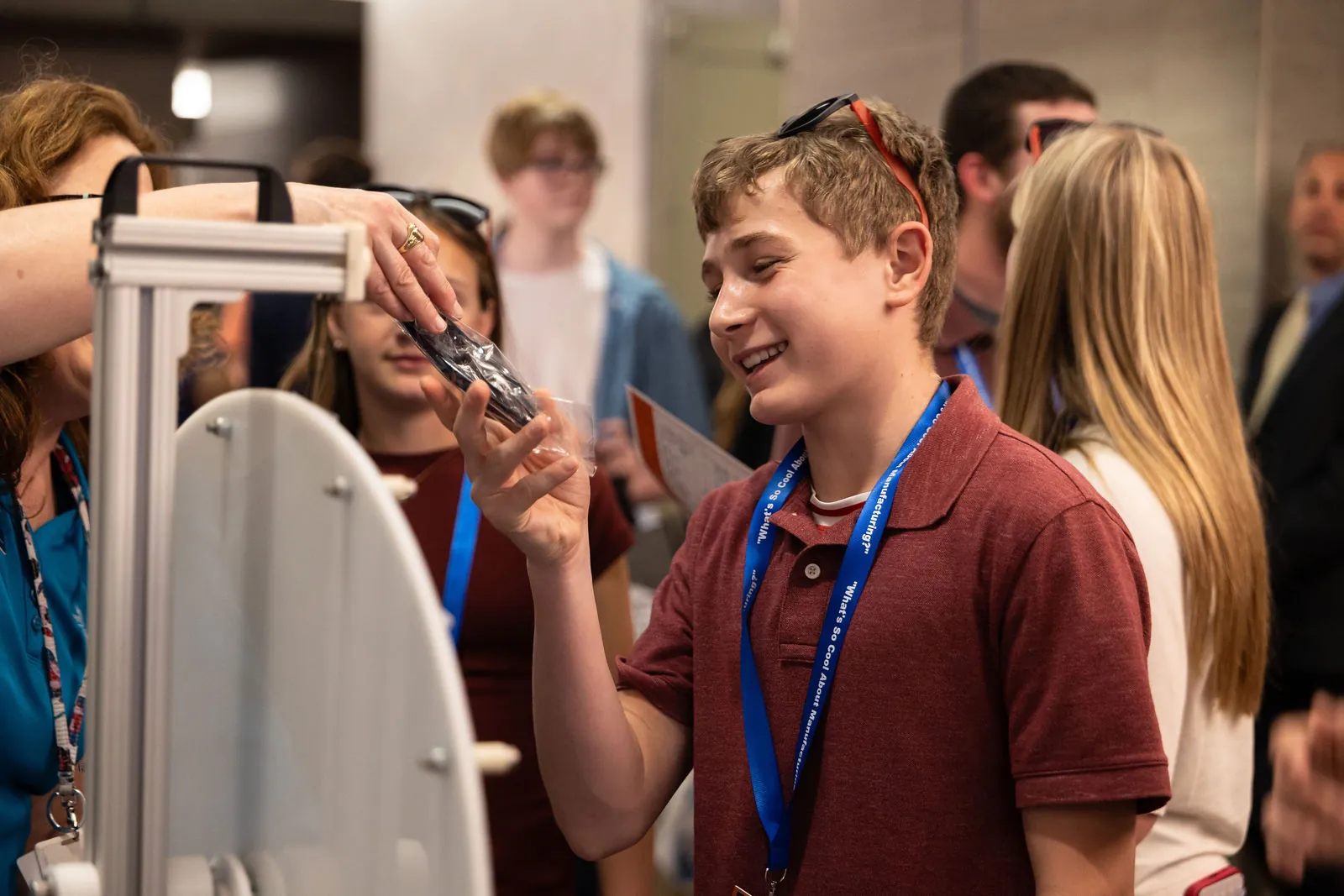 A smiling student in a red polo and lanyard reaching for a giveaway at a vendor table, with other students around him