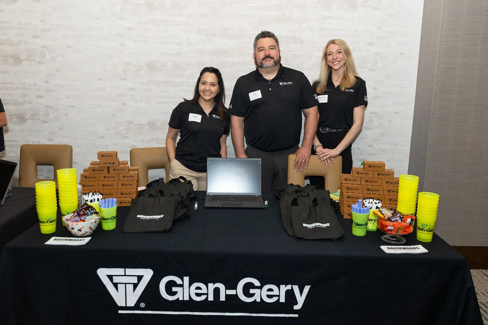 Three people in black shirts behind a Glen-Gery table with a laptop, cups and stacks of small bricks