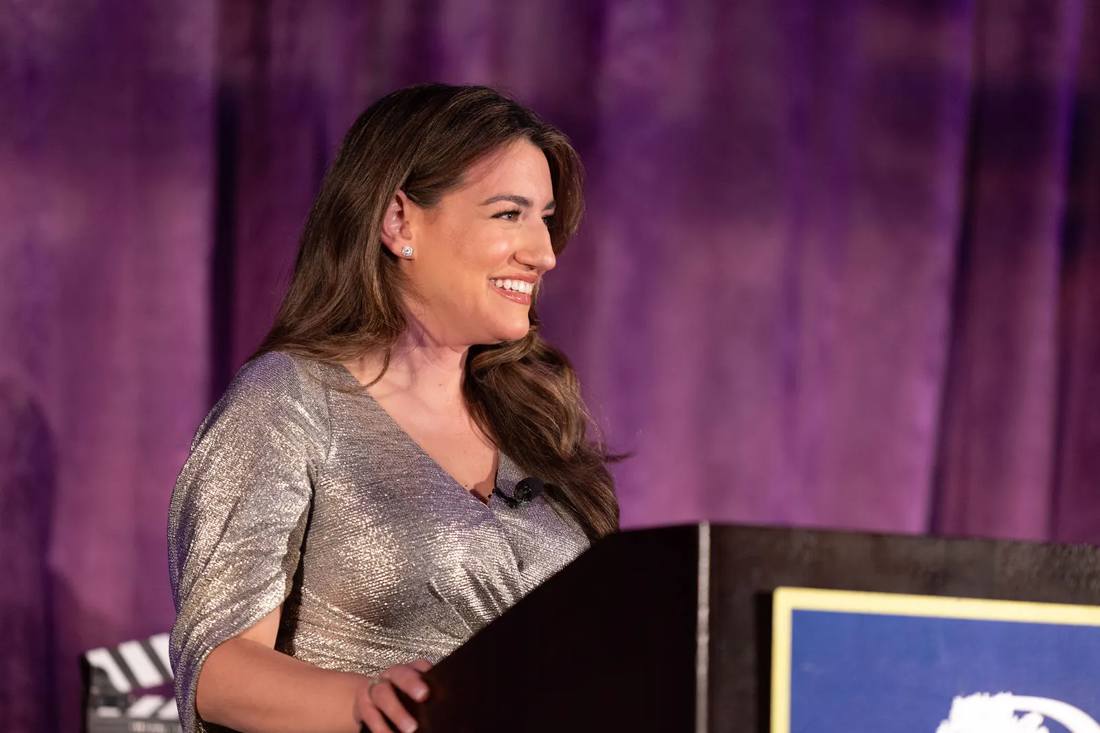 A host in a silver dress smiling at a podium in front of purple curtains
