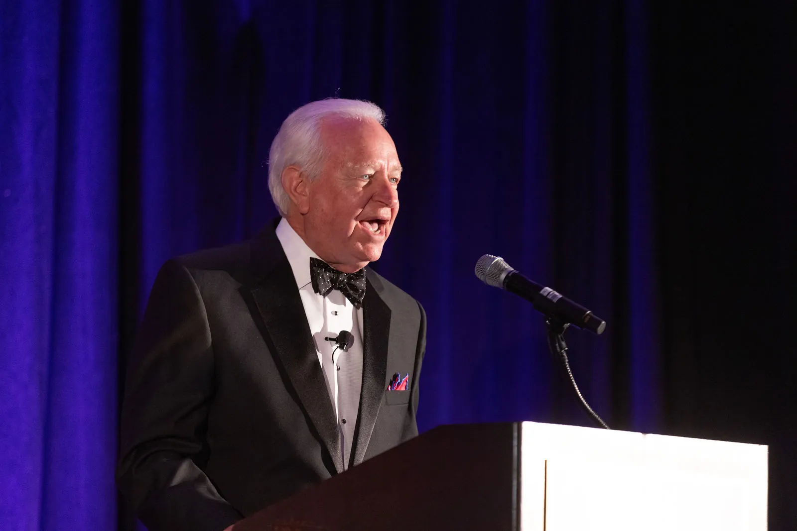 A host in a tuxedo speaking at a podium in front of blue curtains