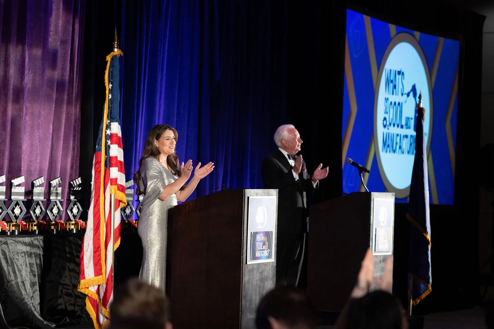 Both hosts applauding on stage beside an American flag, with the contest logo on the screen behind them