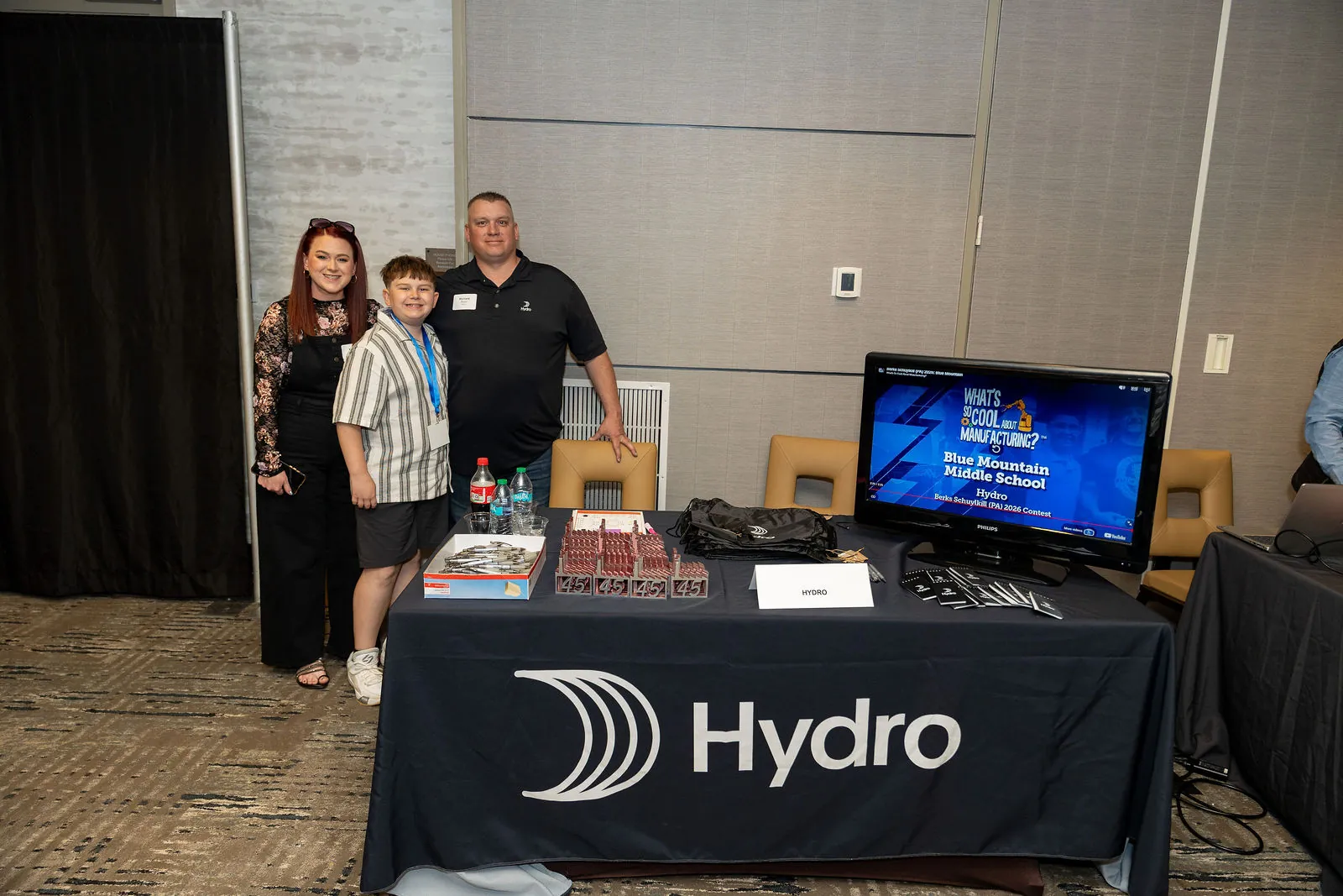 Two adults and a student standing behind the Hydro table, with a screen showing Blue Mountain Middle School