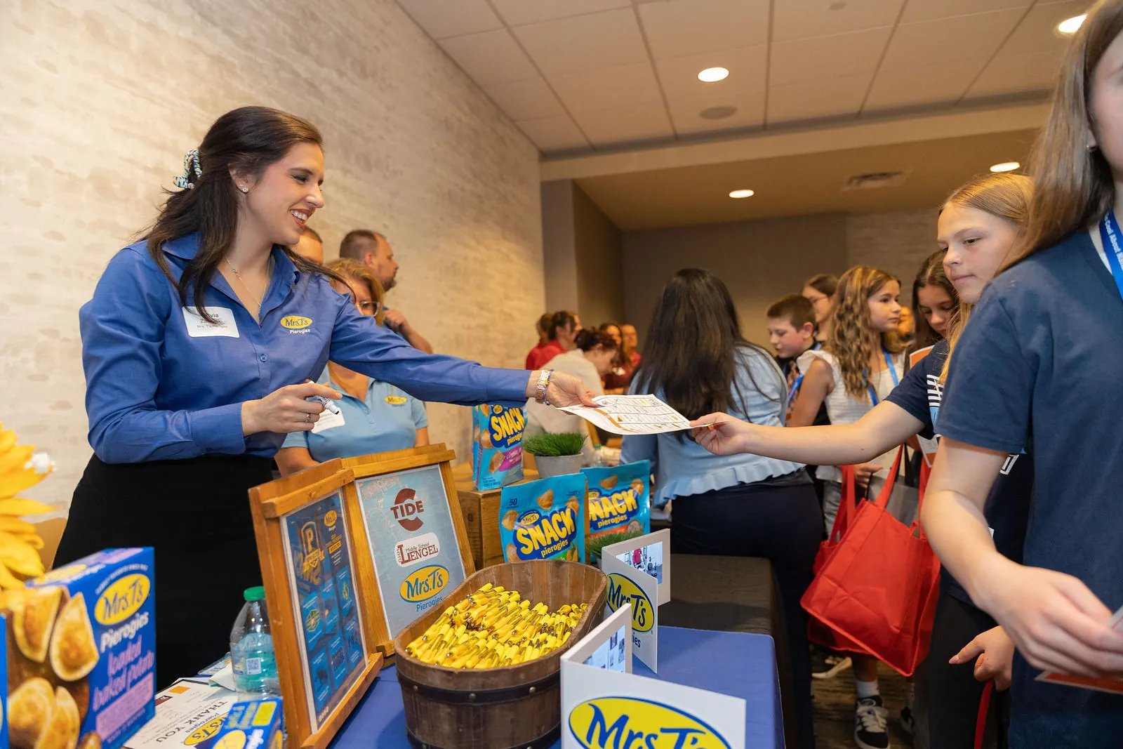 A woman in a blue Mrs. T's shirt handing a card to a student across a table of pierogi snacks and giveaways