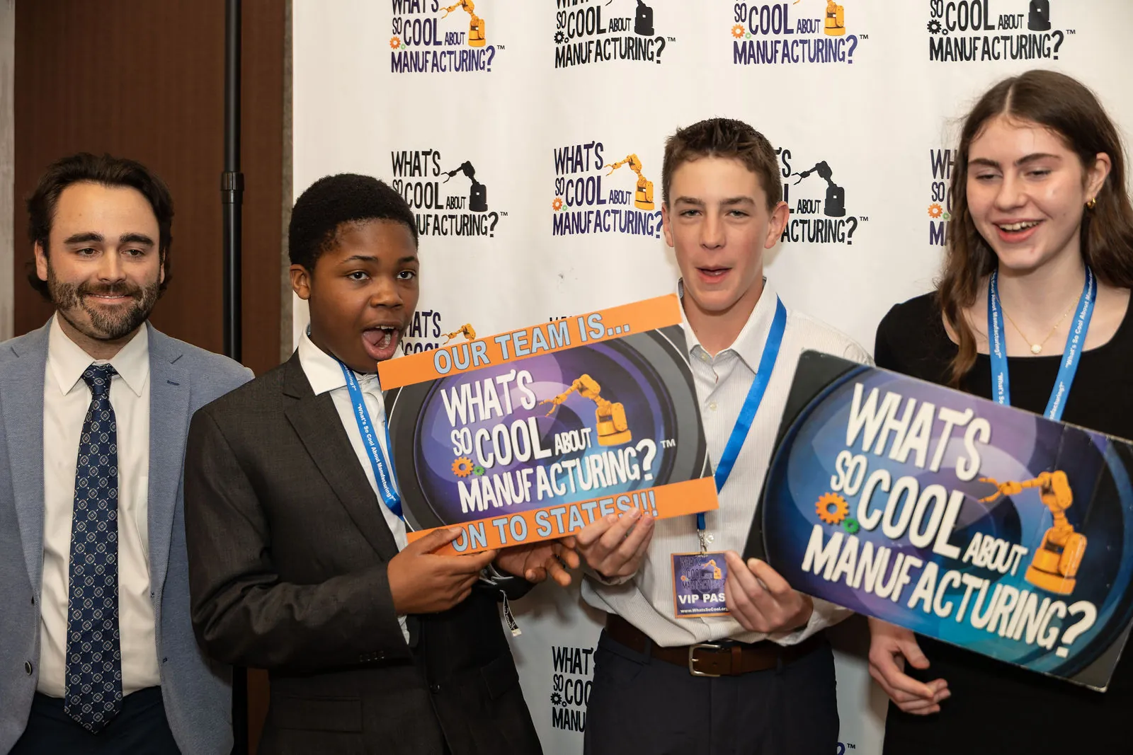 Students and their teacher at the step and repeat holding a What's So Cool About Manufacturing sign and an Our Team Is On To States sign