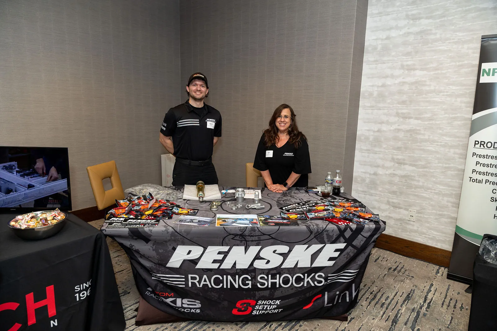 Two people behind a Penske Racing Shocks table covered in giveaways