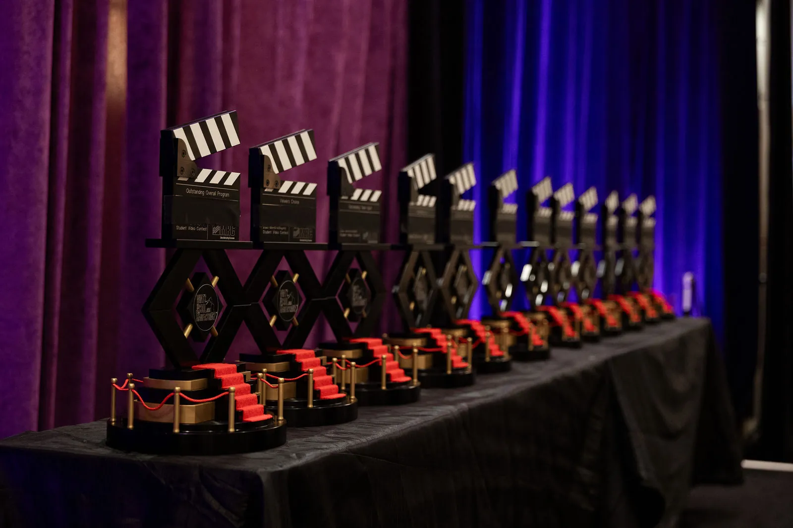 A long row of black clapperboard trophies on a table in front of purple stage curtains